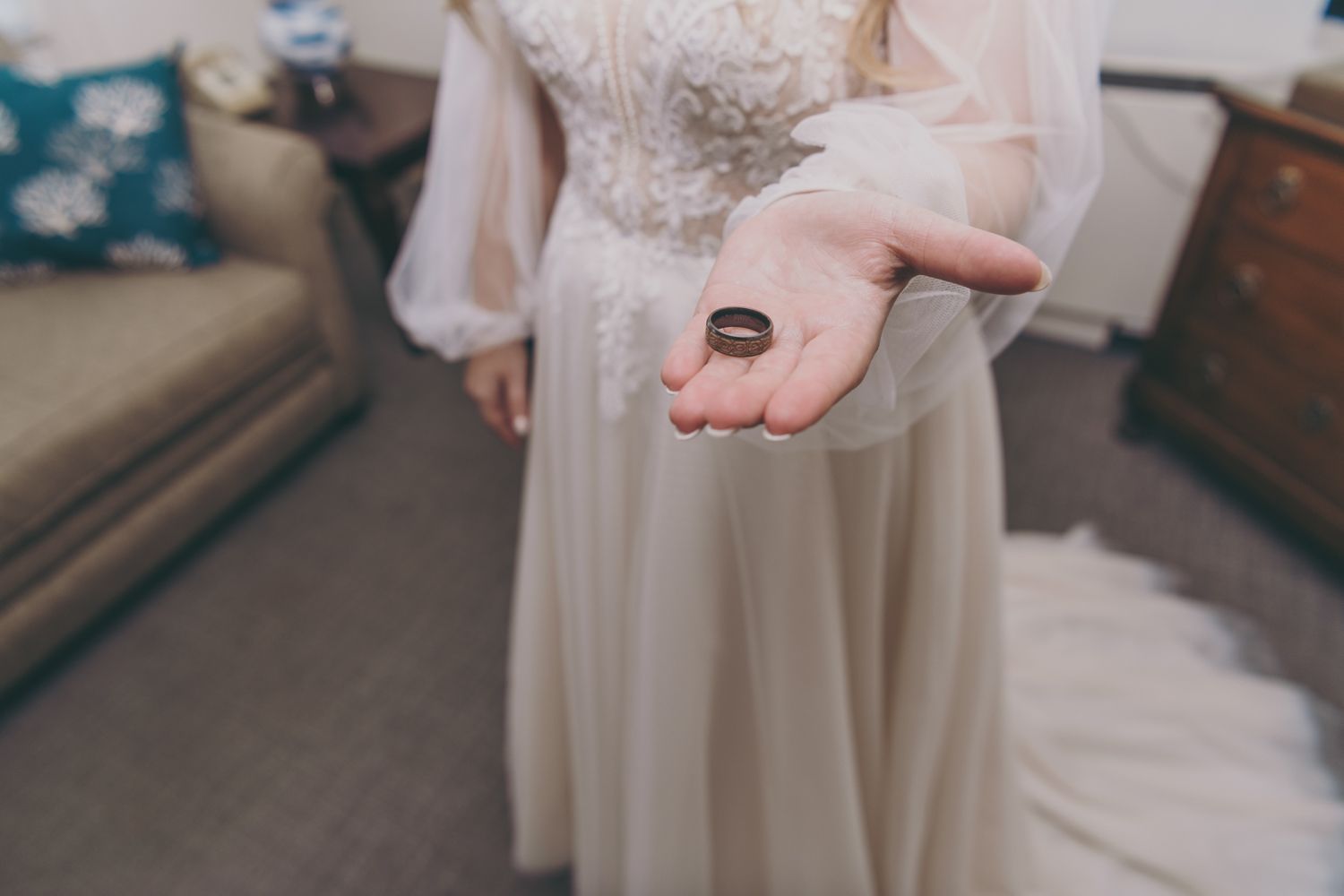 Bride in white lace wedding gown holding a dark wooden wedding band in her outstretched palm.