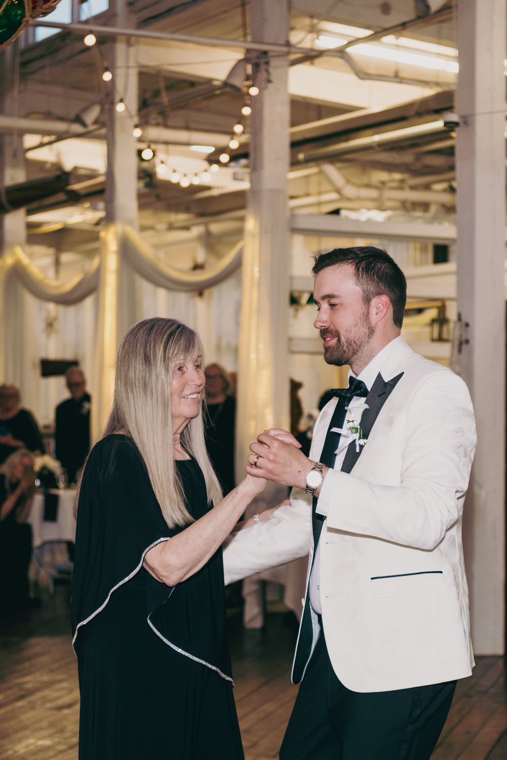 Groom in white tuxedo shares mother-son dance on wooden floor at wedding reception in industrial venue.