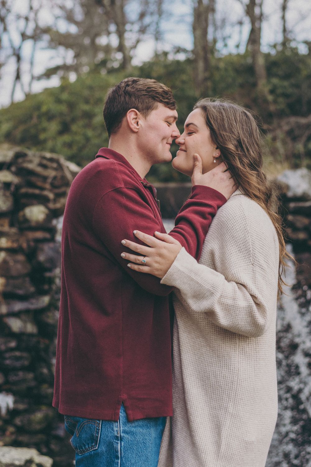 A couple shares a romantic moment outdoors, nearly kissing, dressed in cozy sweaters near a stone wall.