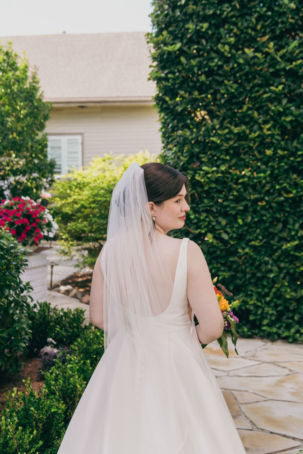 Bride in elegant backless white gown and veil stands in a lush garden holding a colorful bridal bouquet.
