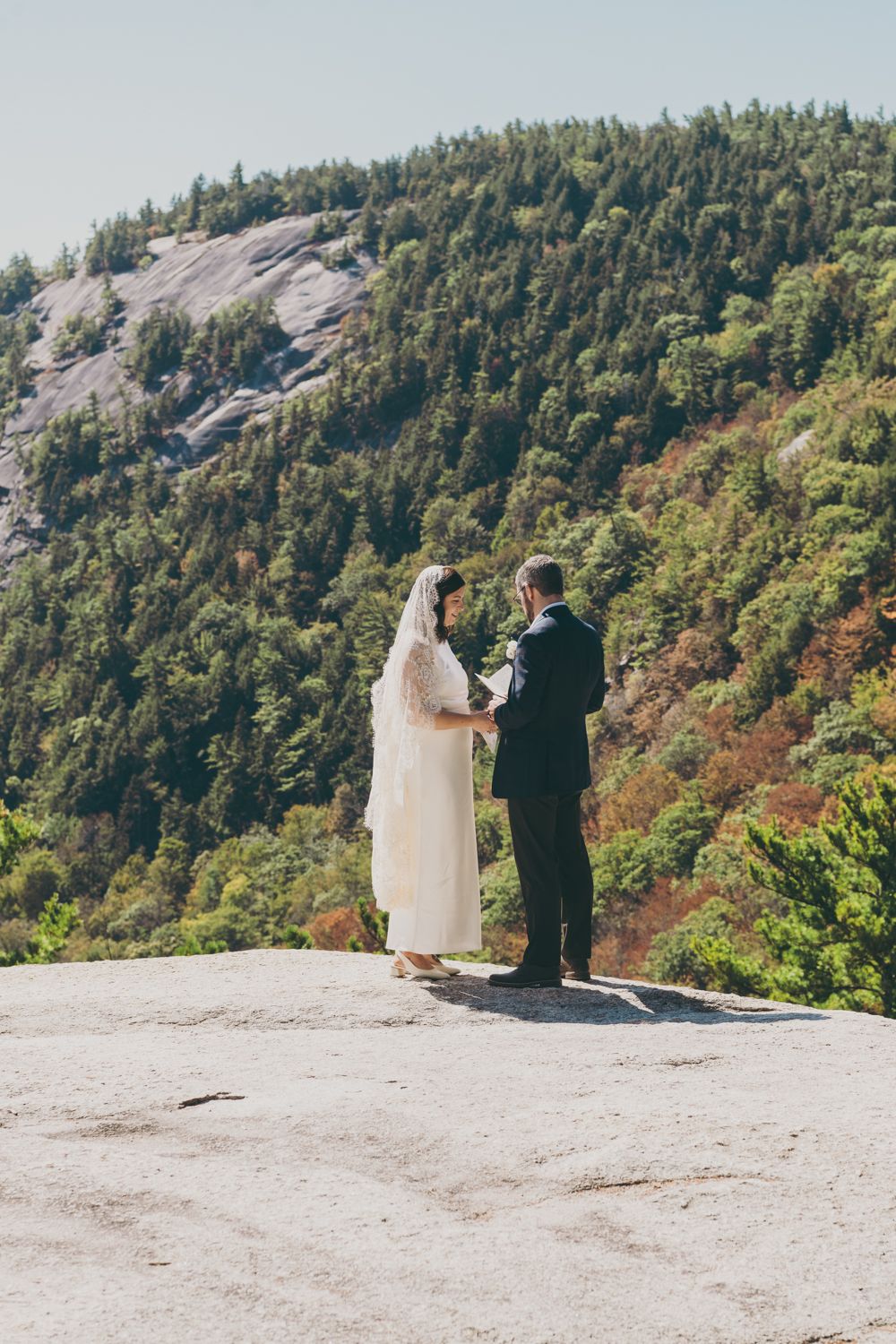 Bride and groom exchanging vows on granite mountain summit with forested hillside backdrop.