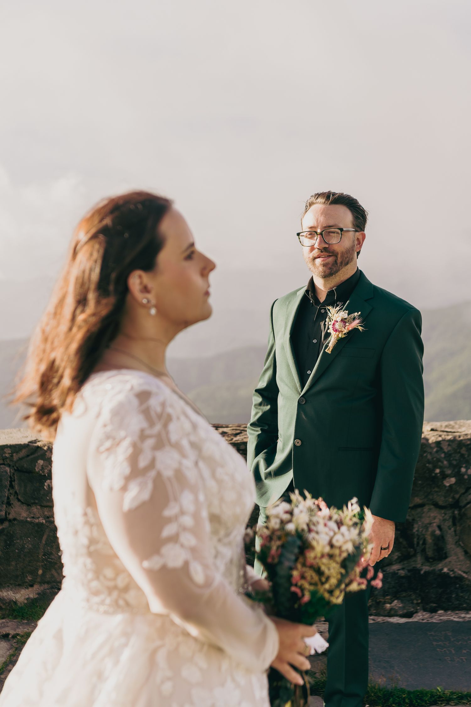 Bride in lace gown holds bouquet as groom in green suit smiles at her on a scenic mountain overlook.