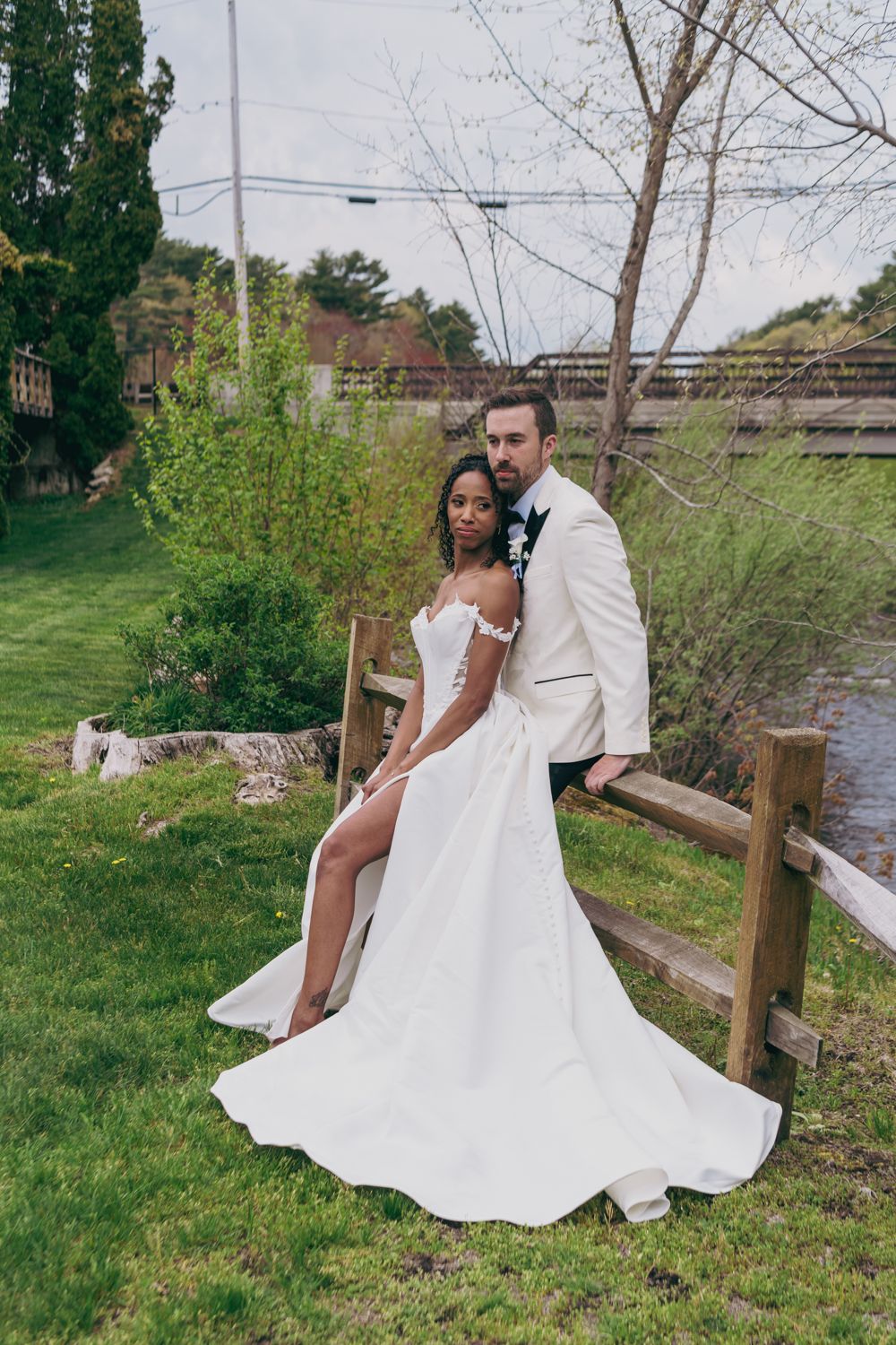 Elegant interracial bride and groom posing by a wooden fence in a garden, bride in a slit wedding gown.