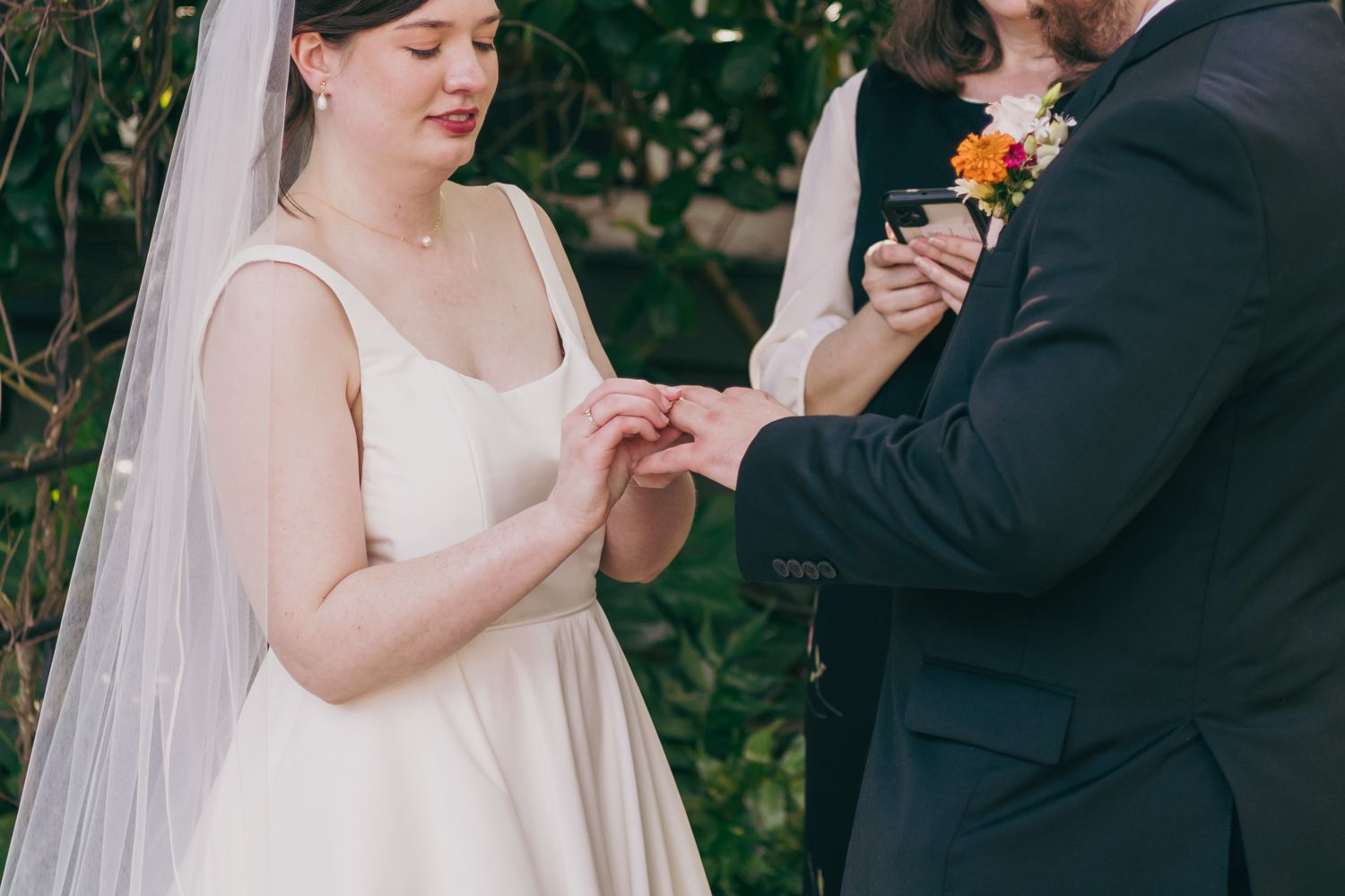 Bride places wedding ring on grooms finger during outdoor garden wedding ceremony.
