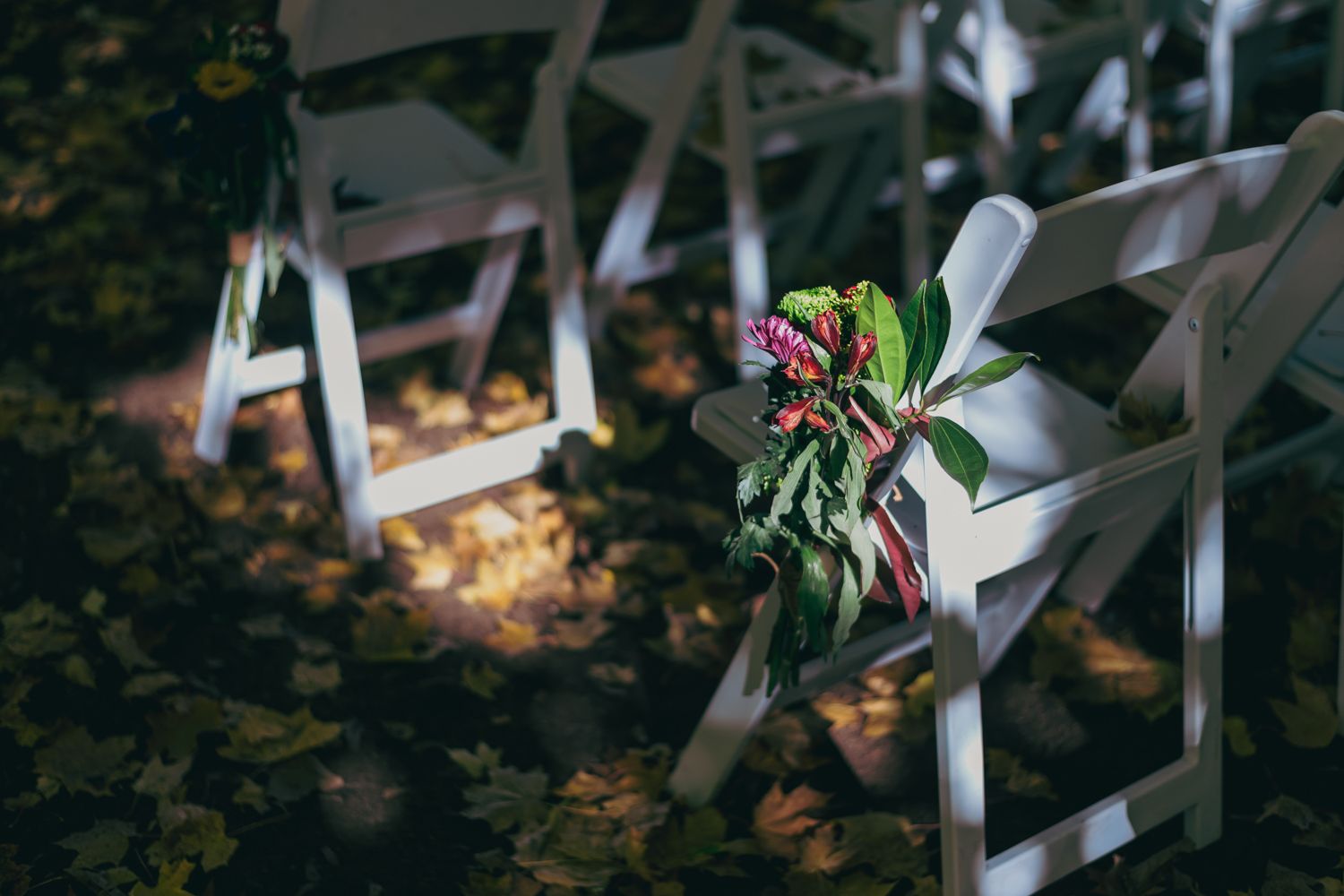 White folding wedding ceremony chairs adorned with floral greenery bouquets on autumn leaf-covered ground.