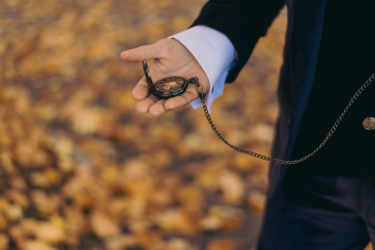 Groom holding vintage pocket watch on chain against autumn leaf background at wedding.