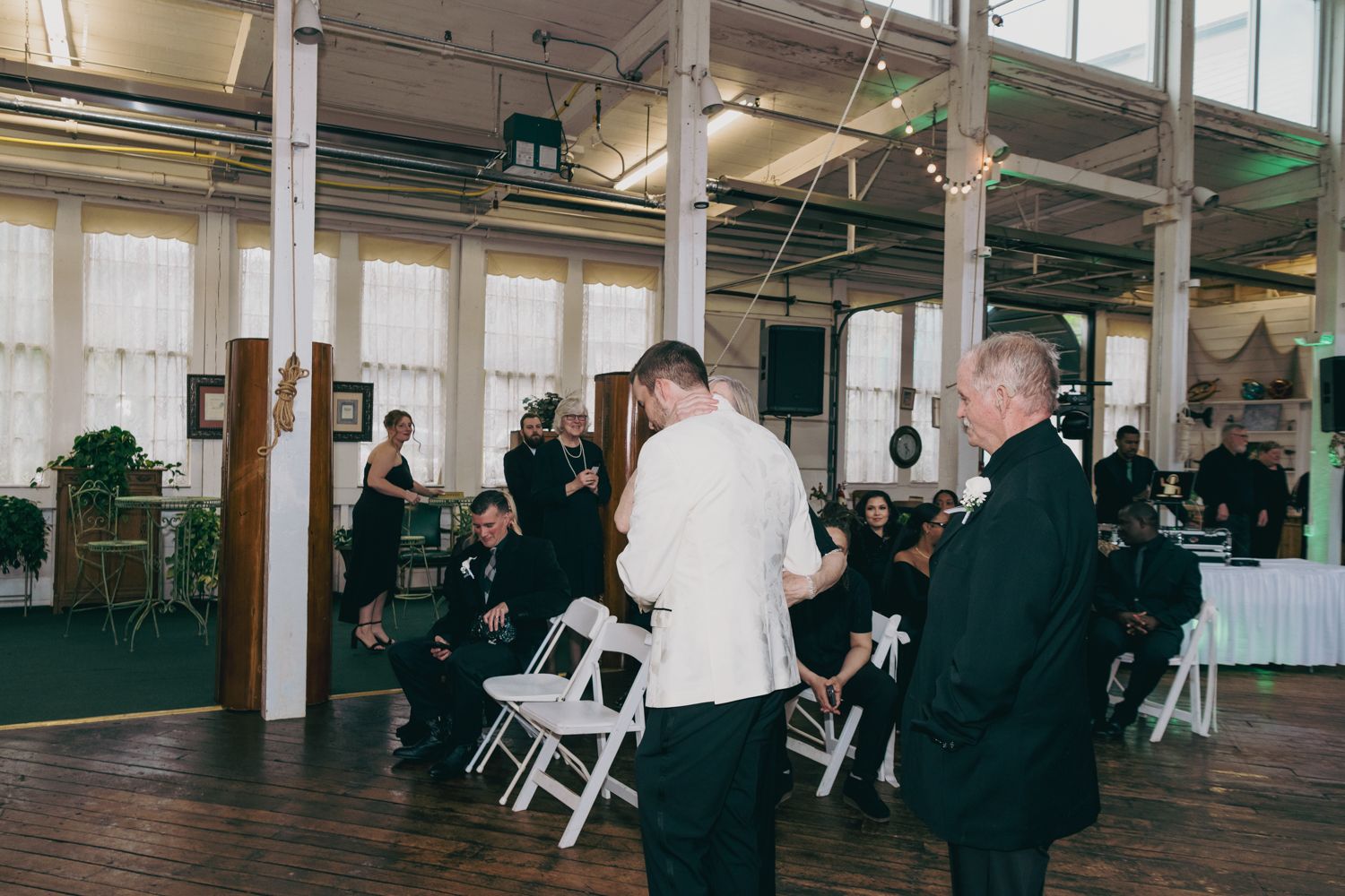 Guests mingle in an elegant industrial wedding venue with white folding chairs and string lights overhead.