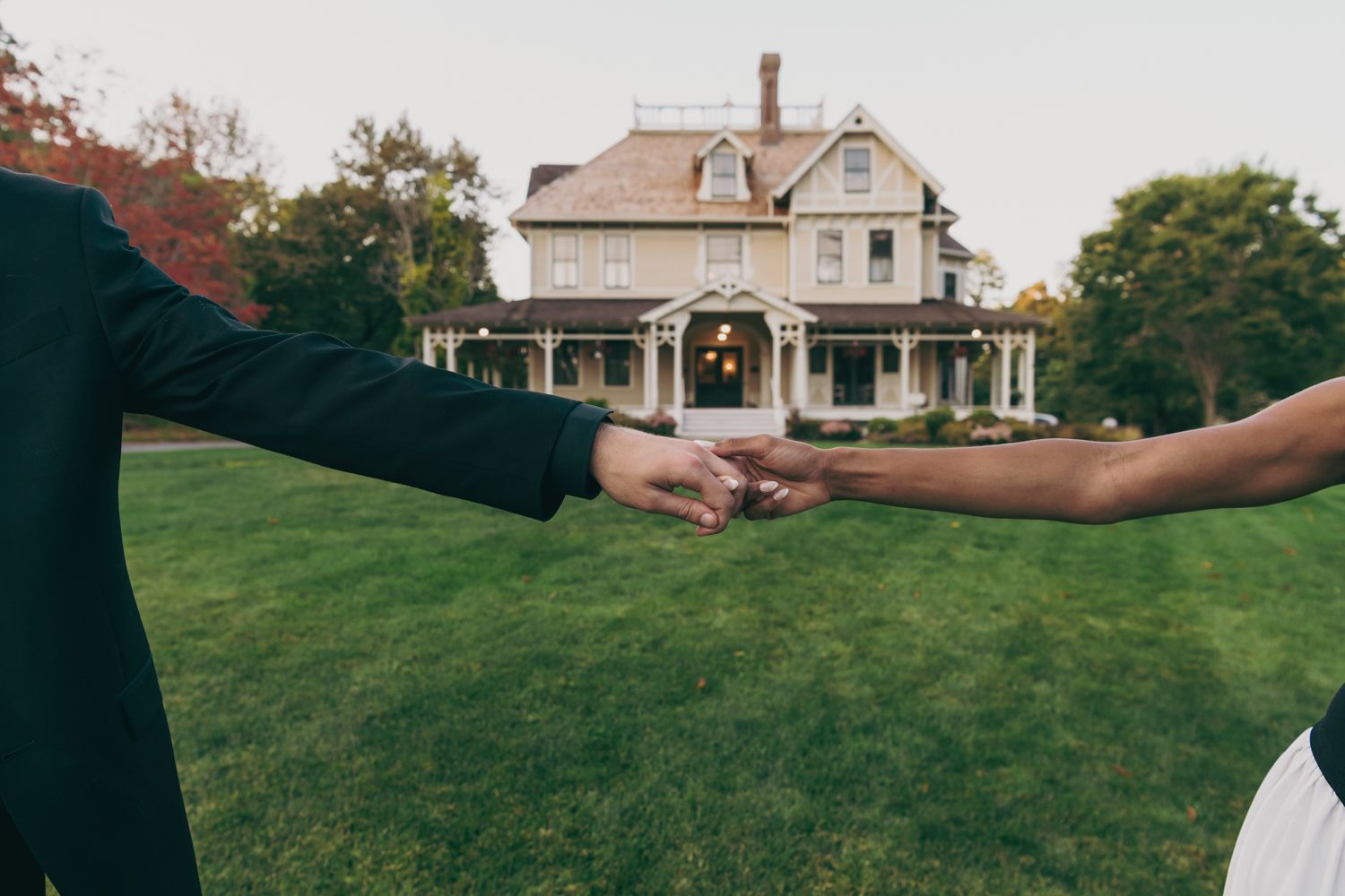 Couple holding hands on a green lawn in front of a charming Victorian house during autumn.