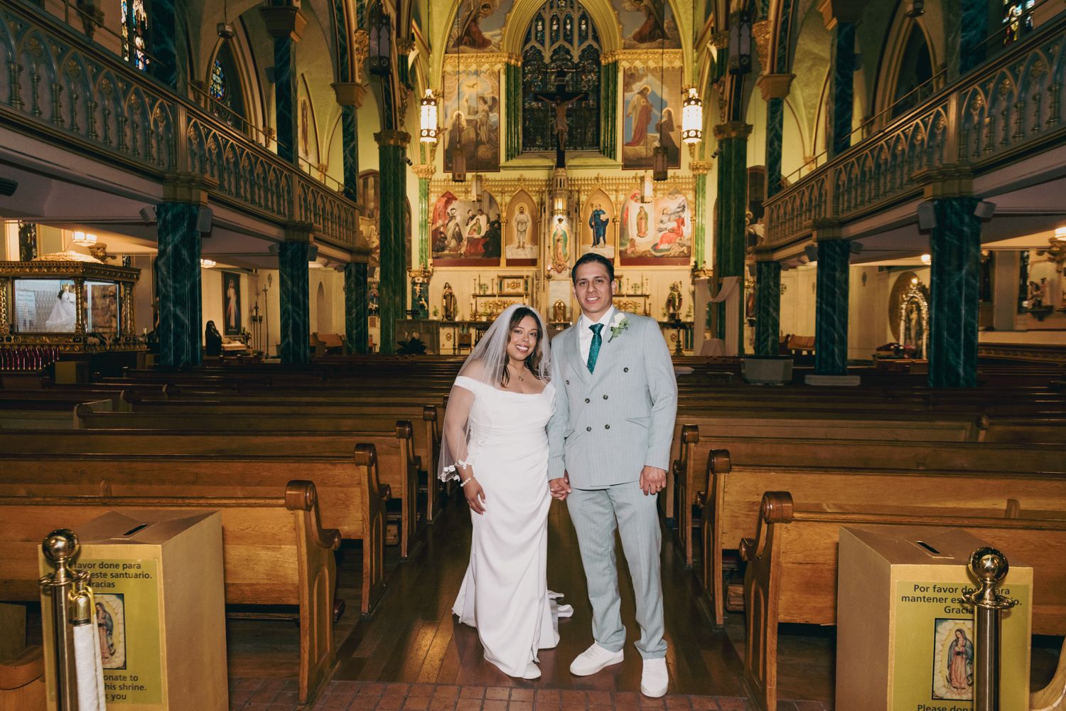 Bride and groom posing together inside an ornate church with wooden pews and colorful stained glass windows.
