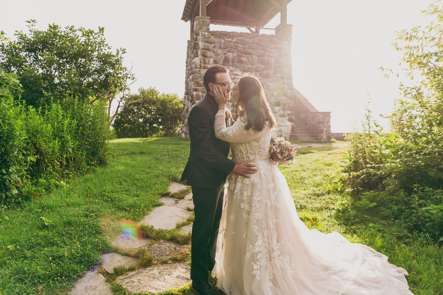 Bride and groom share a kiss at golden hour near a rustic stone tower surrounded by lush greenery.