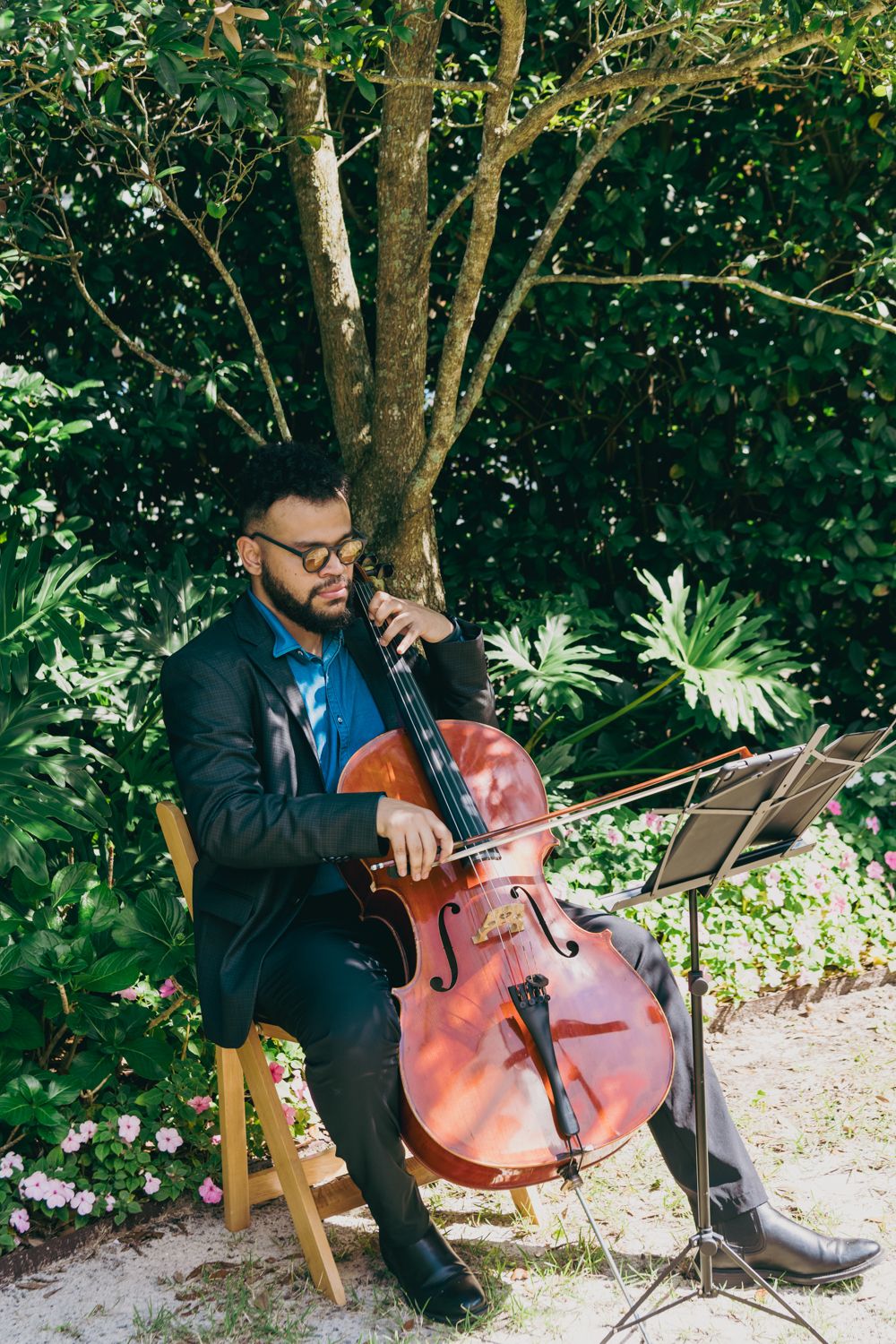 Musician in a black suit plays cello outdoors at a garden wedding ceremony.