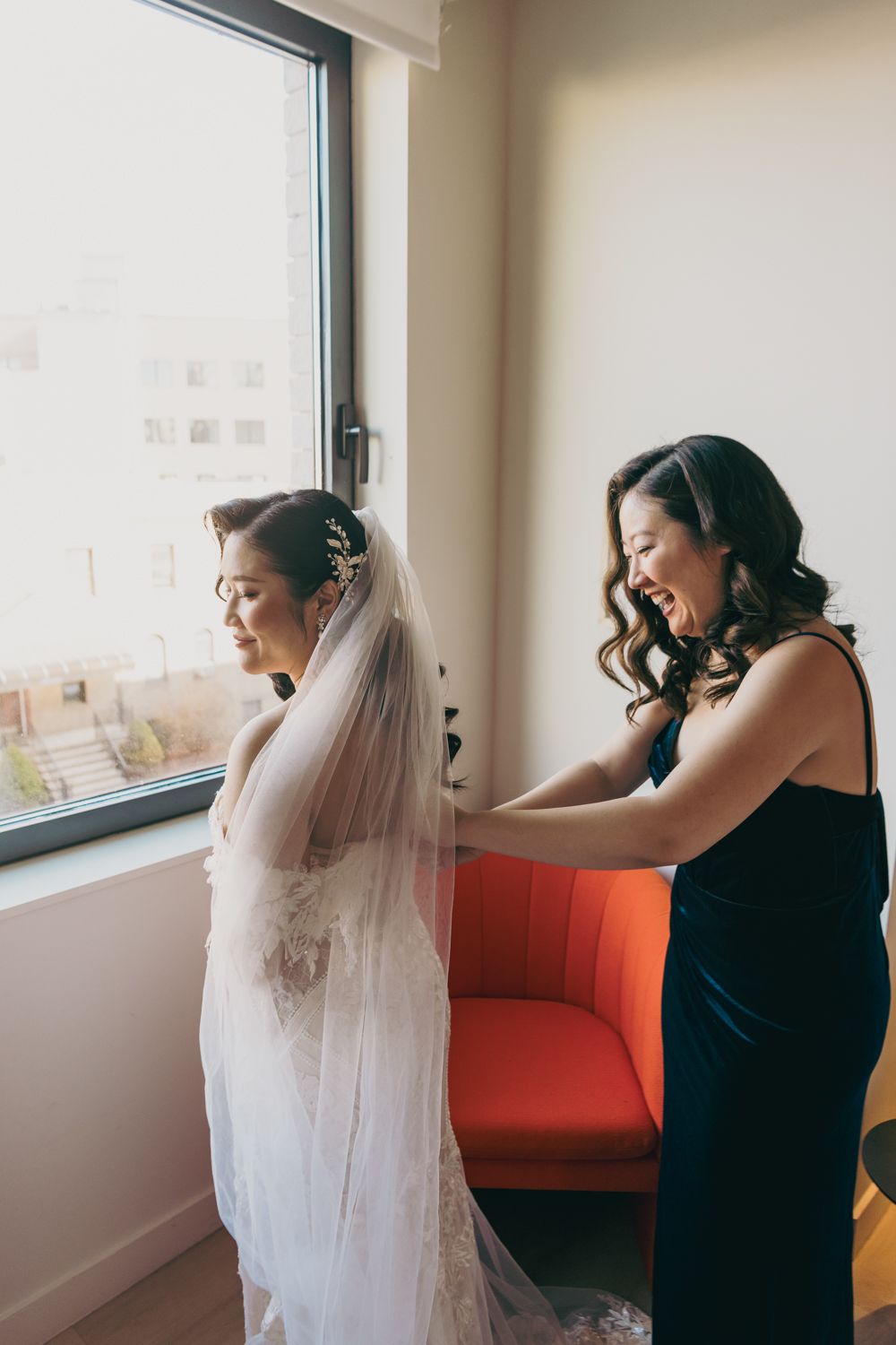 A smiling bridesmaid in navy blue helps a bride in a white gown and veil prepare near a sunlit window.