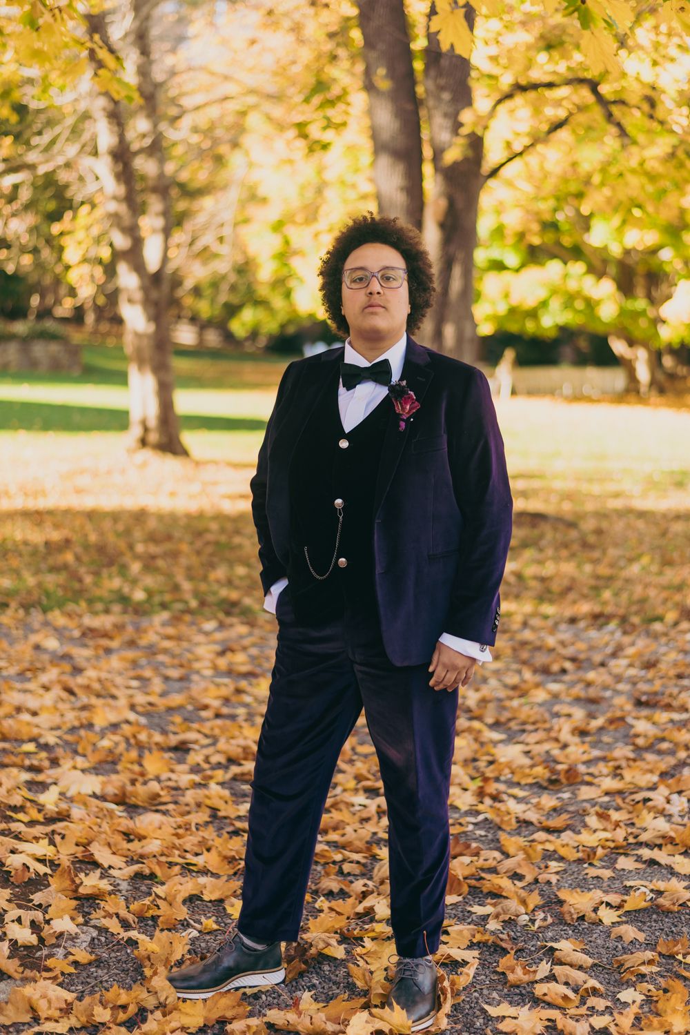 Groom in navy velvet tuxedo with bow tie standing among golden autumn leaves.