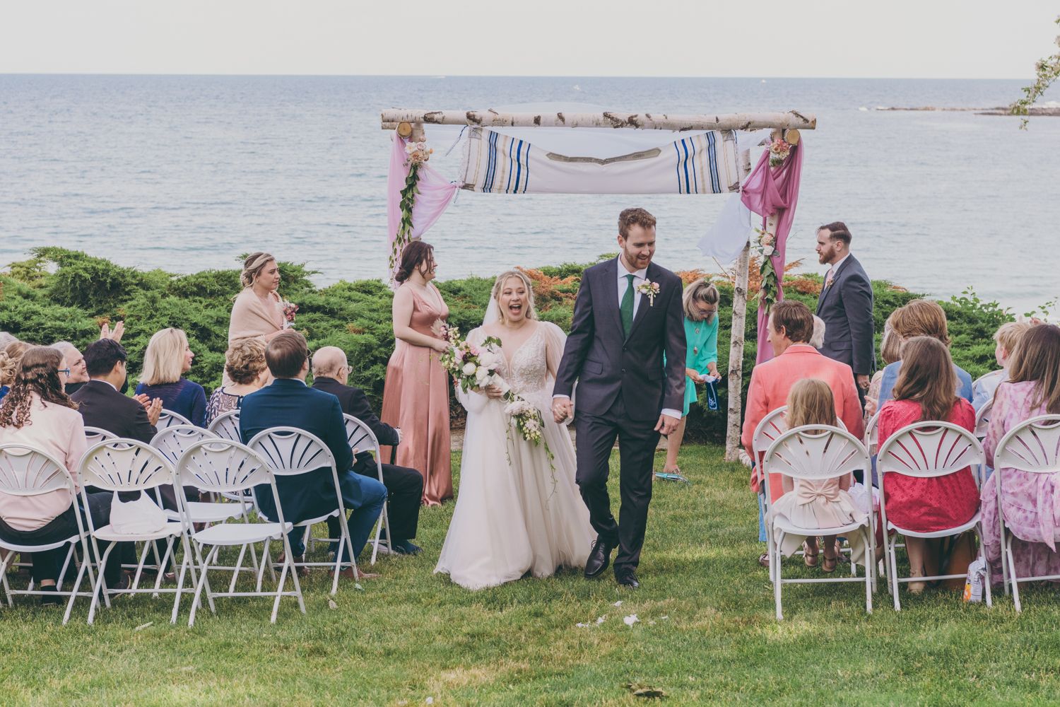 Newlyweds walk down the aisle hand in hand after outdoor seaside wedding ceremony with seated guests.