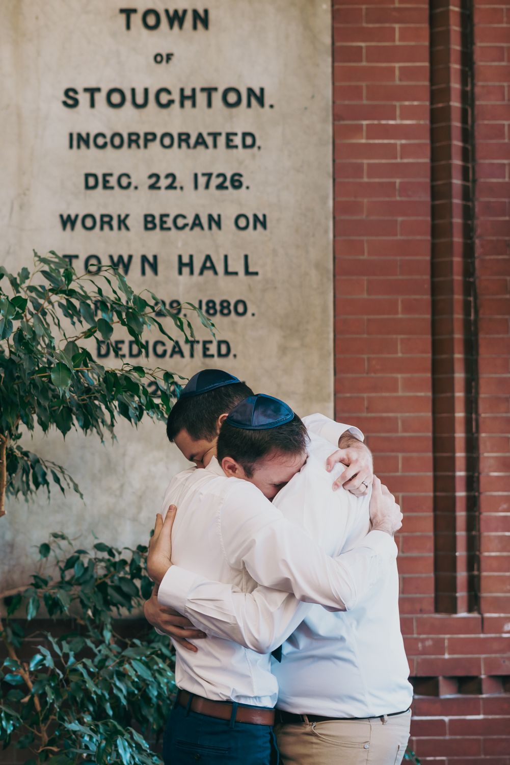 Two men in white shirts and yarmulkes share an emotional embrace outside Stoughton Town Hall, incorporated 1726.