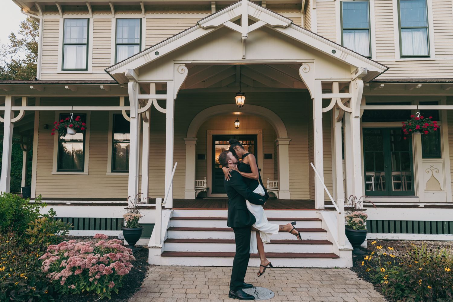 Romantic interracial couple embracing on steps of Victorian home with wrap-around porch and floral gardens.