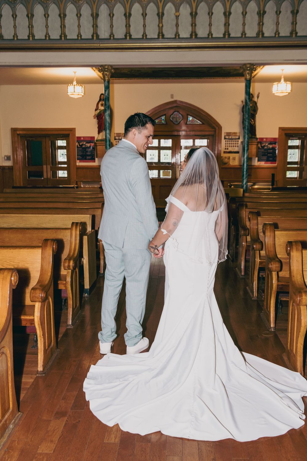 Bride and groom holding hands in church aisle, bride wearing cathedral veil and fitted gown.