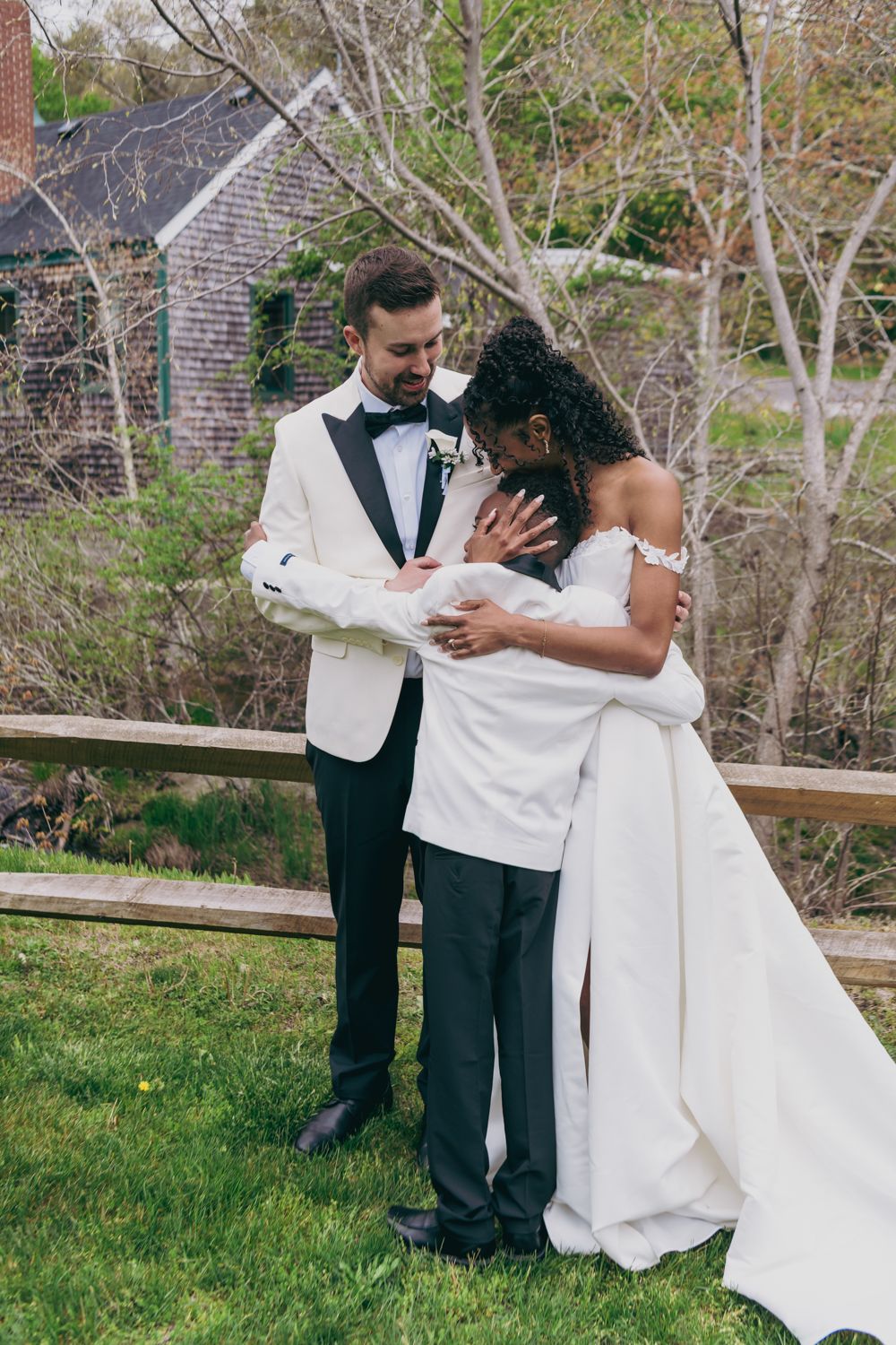 Interracial newlyweds embracing tenderly outdoors by a wooden fence after wedding ceremony.