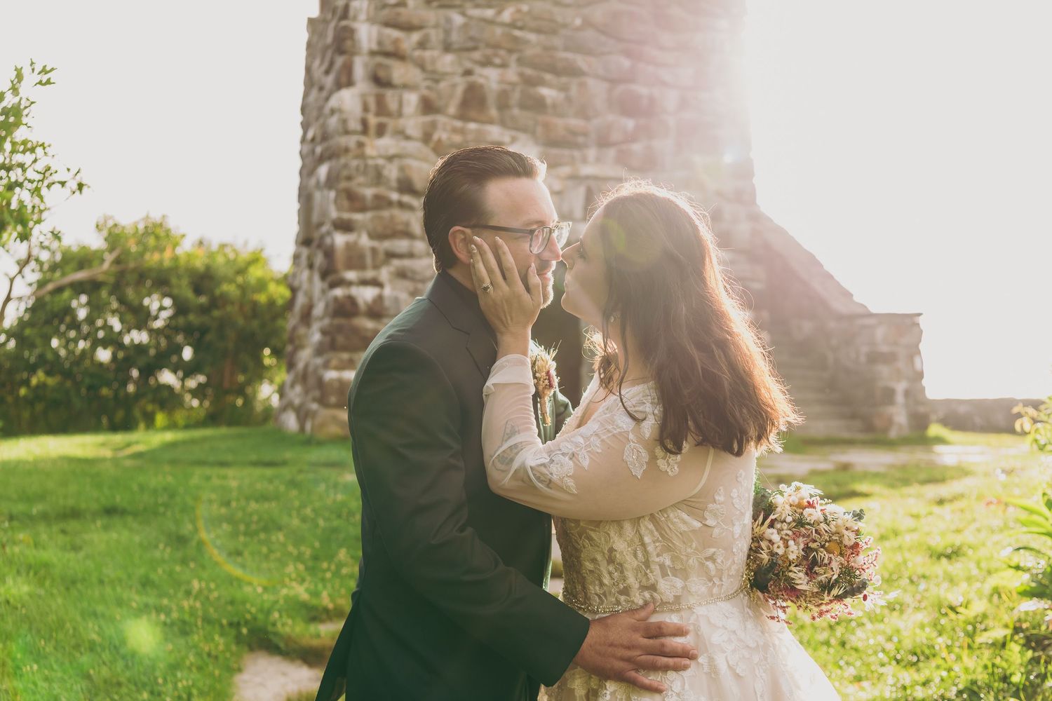Bride and groom share a romantic moment outdoors near a stone tower, bathed in golden sunlight on their wedding day.