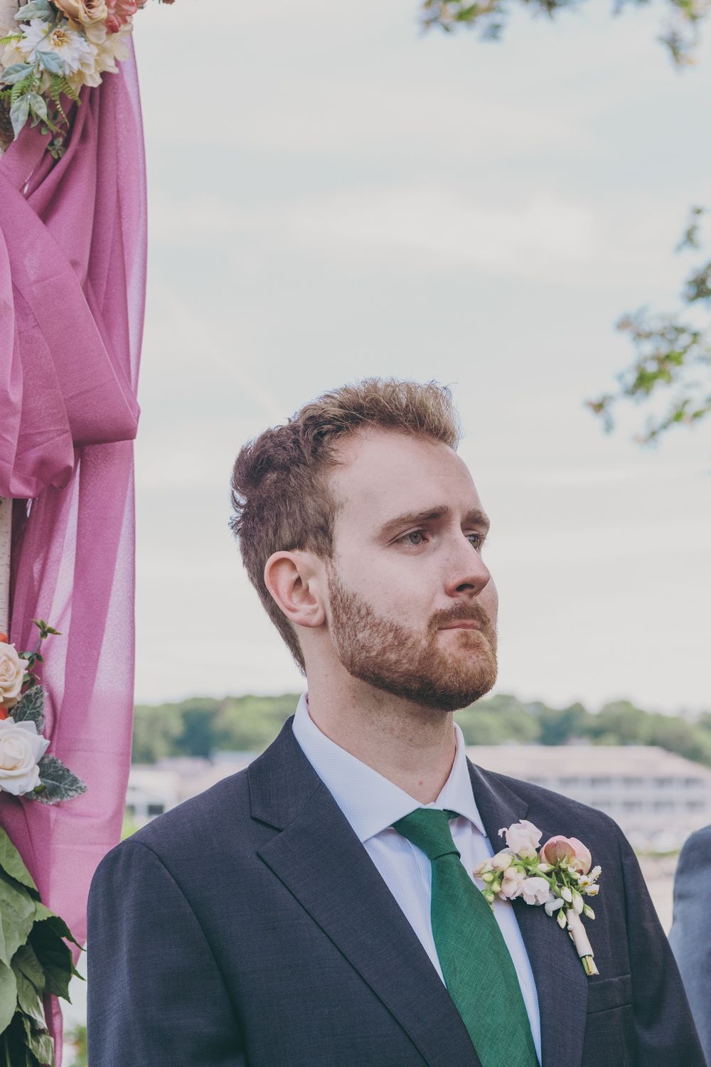 A bearded groom in a navy suit and green tie stands at the altar with a floral boutonniere.