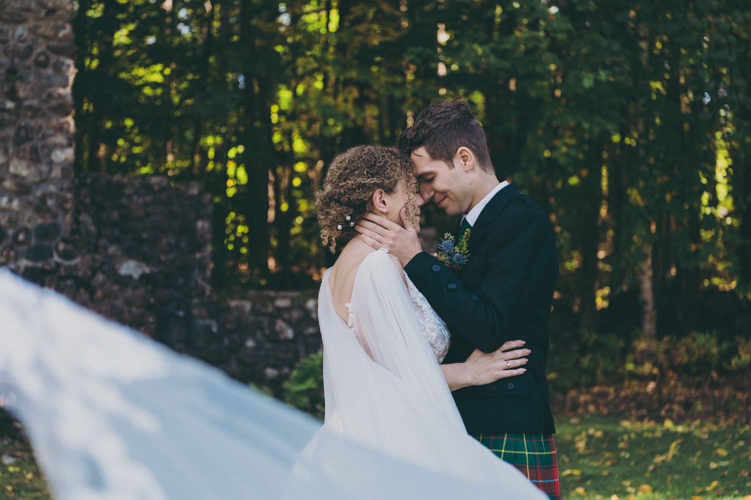 A bride and groom share a romantic moment in a lush green forest, with her white veil flowing in the wind.
