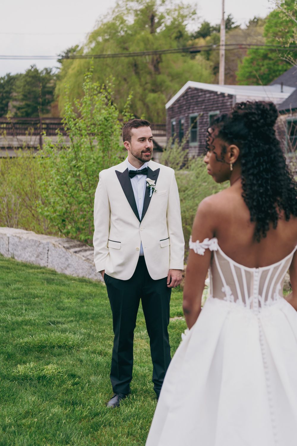 Groom in white tuxedo jacket gazes at bride in strapless ballgown during outdoor first look wedding moment.