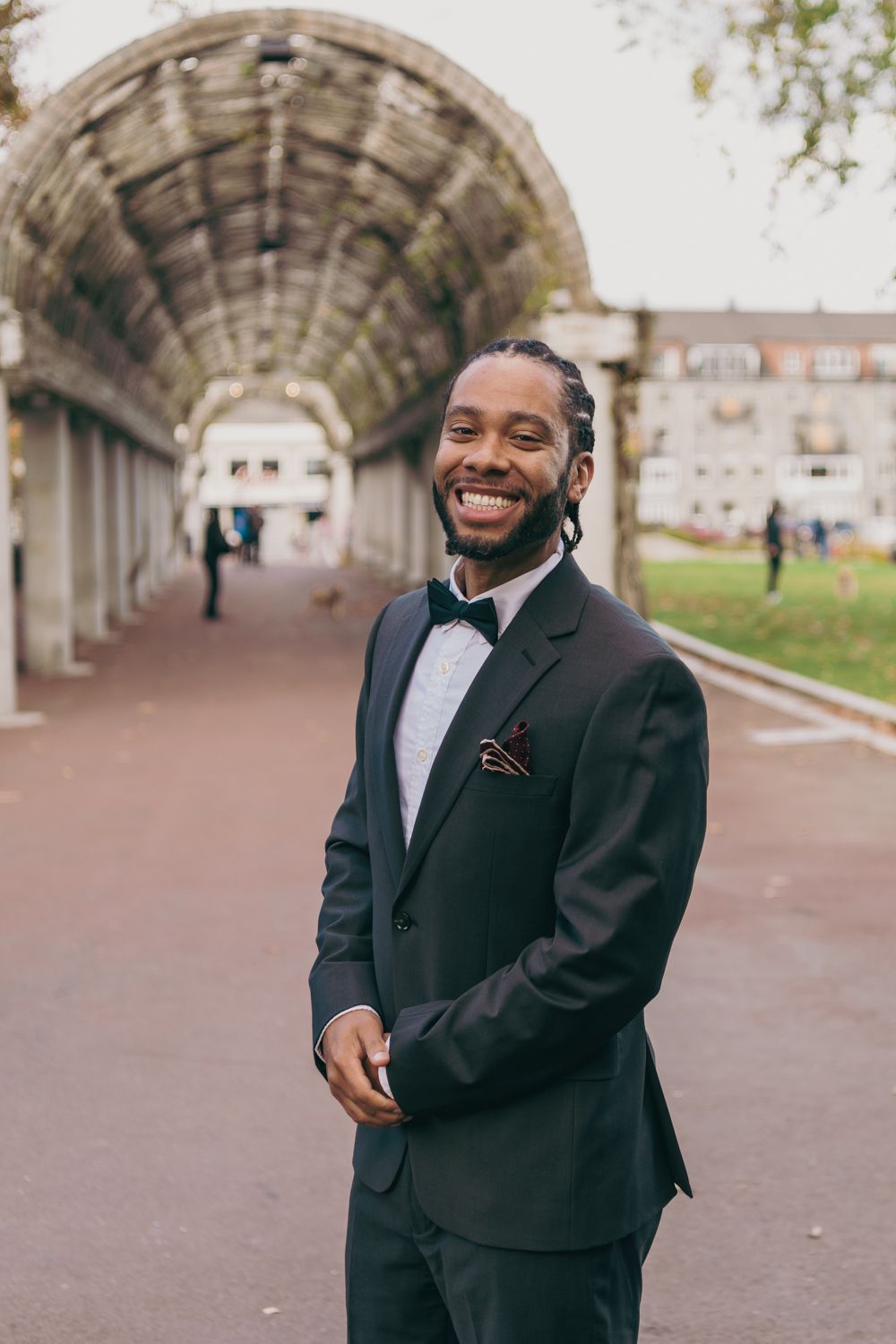 Smiling groom in black tuxedo with bow tie and pocket square posing outdoors under an arched walkway.