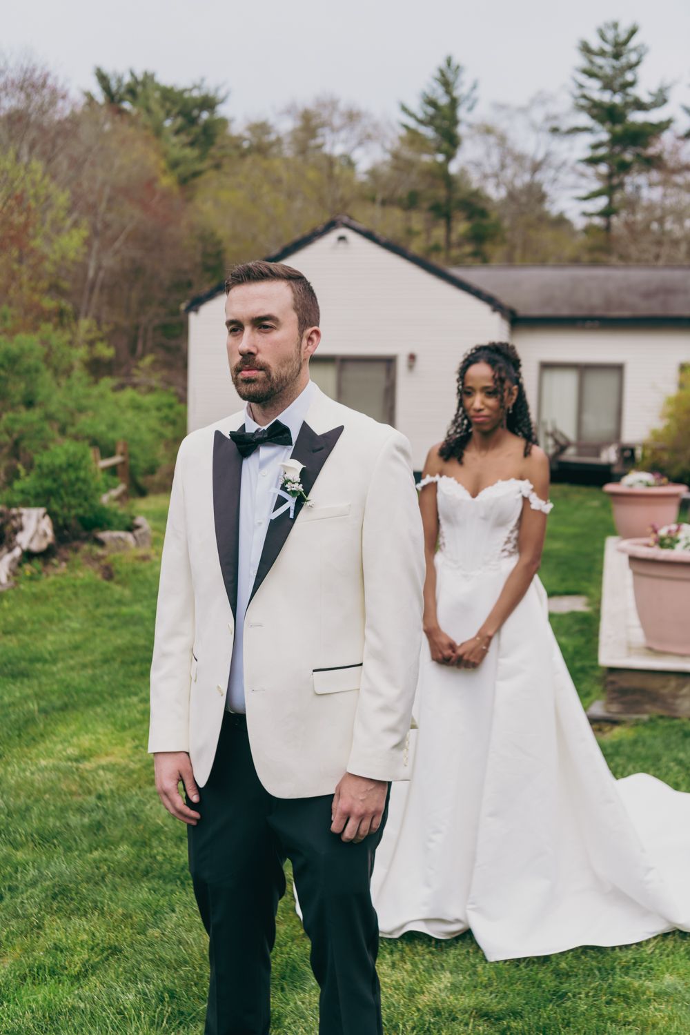 Groom in white tuxedo stands in foreground as bride in strapless gown waits behind him for first look.