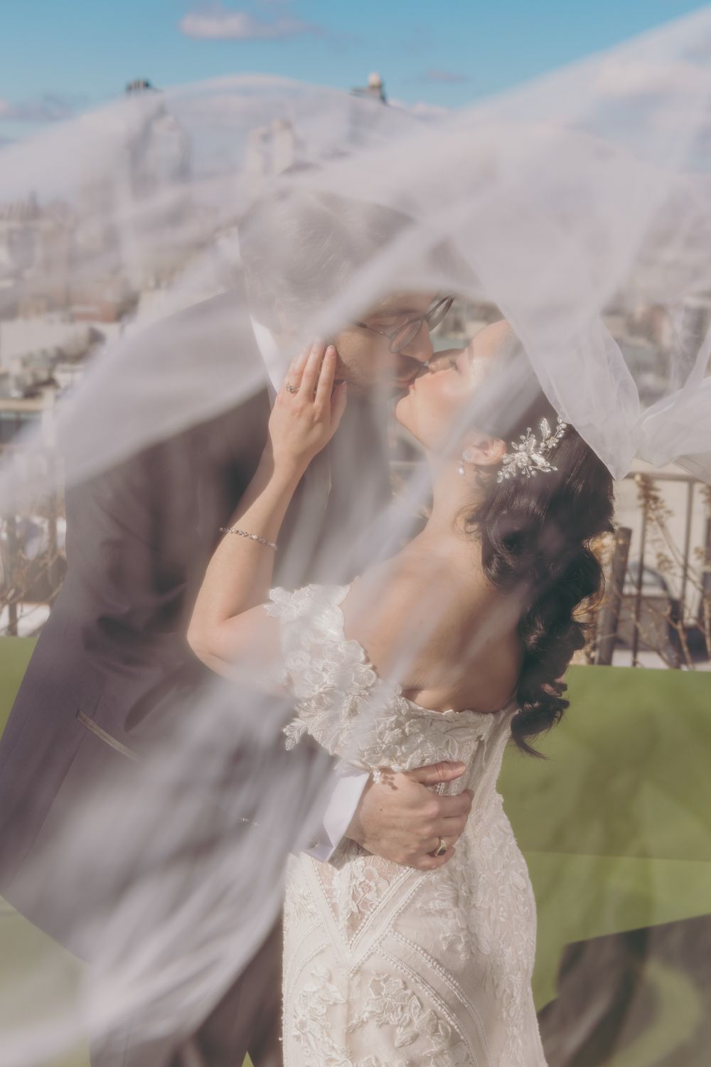 Bride and groom sharing a romantic kiss under a flowing white veil on their wedding day outdoors.