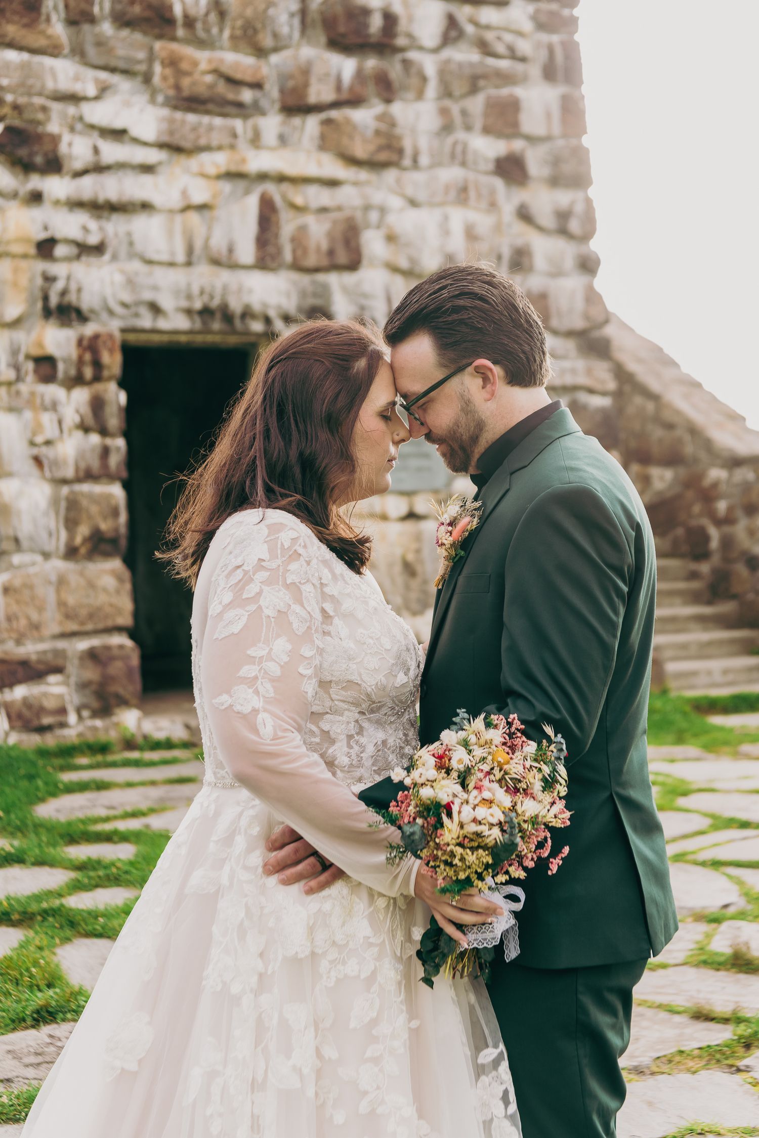 Bride and groom share an intimate moment, touching foreheads while holding a wildflower bouquet before a stone building.