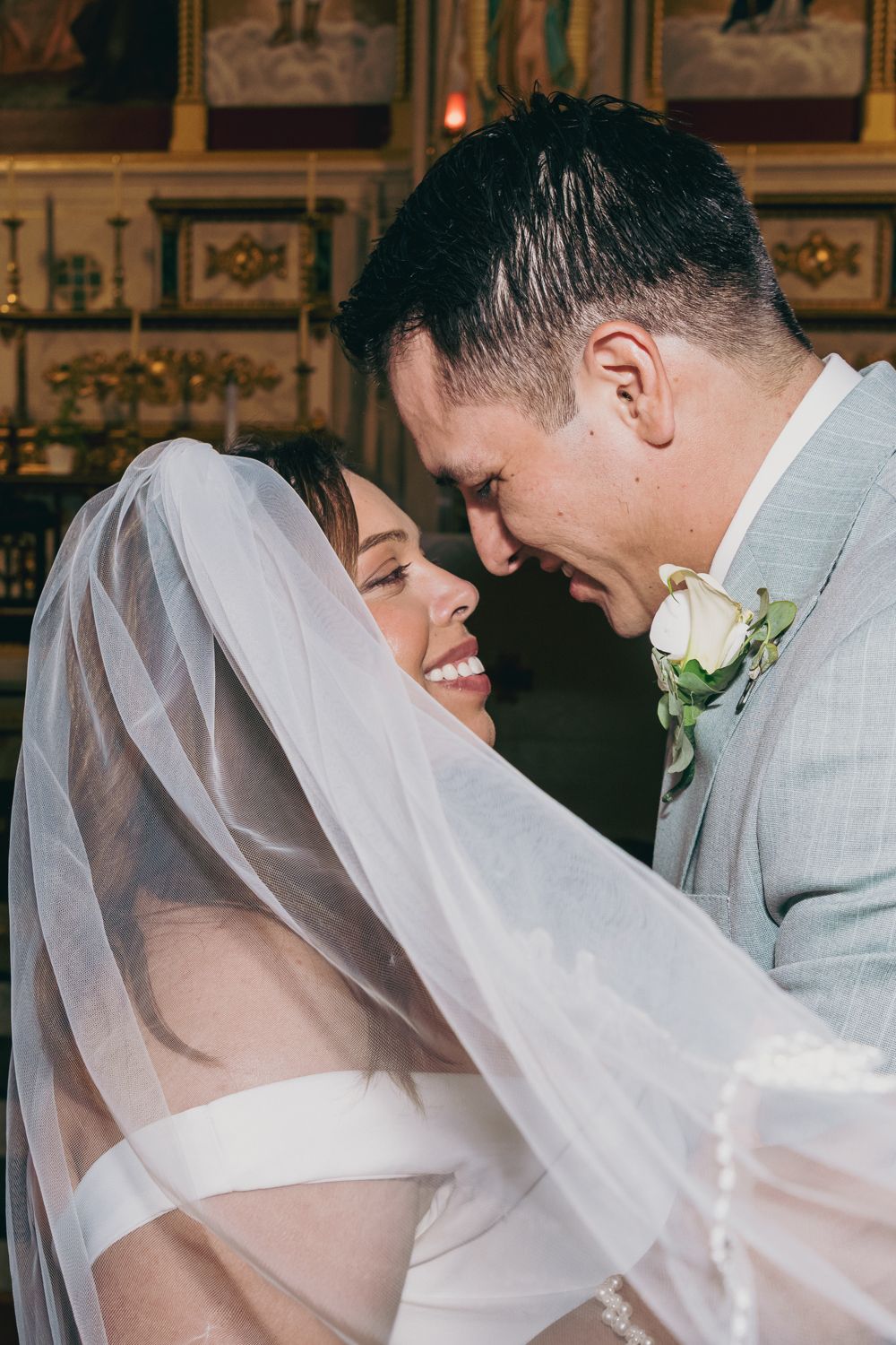 Smiling bride in veil and groom in gray suit with rose boutonniere sharing a tender moment at church altar.