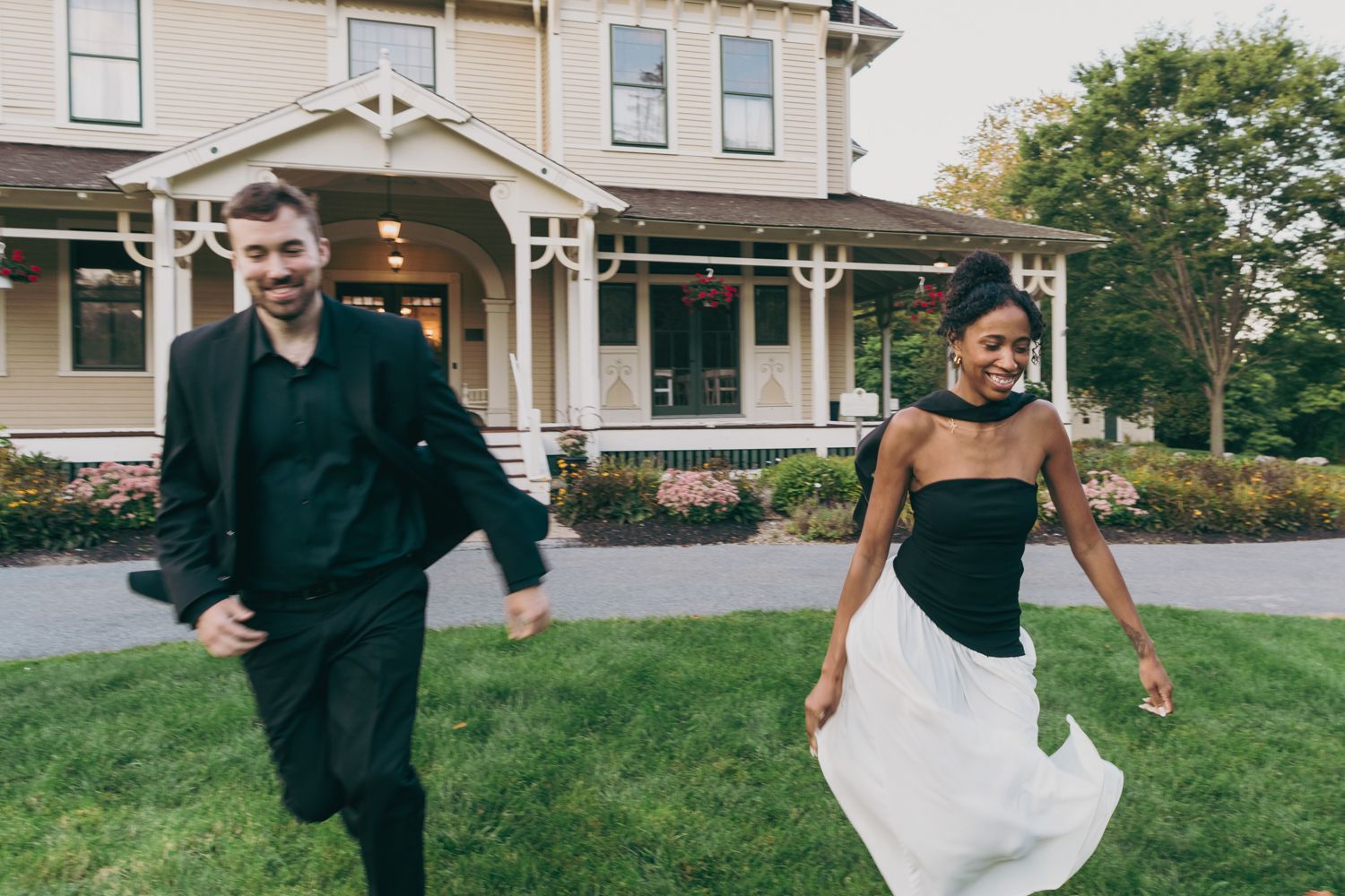 Joyful interracial couple running and laughing on green lawn in front of elegant Victorian mansion.