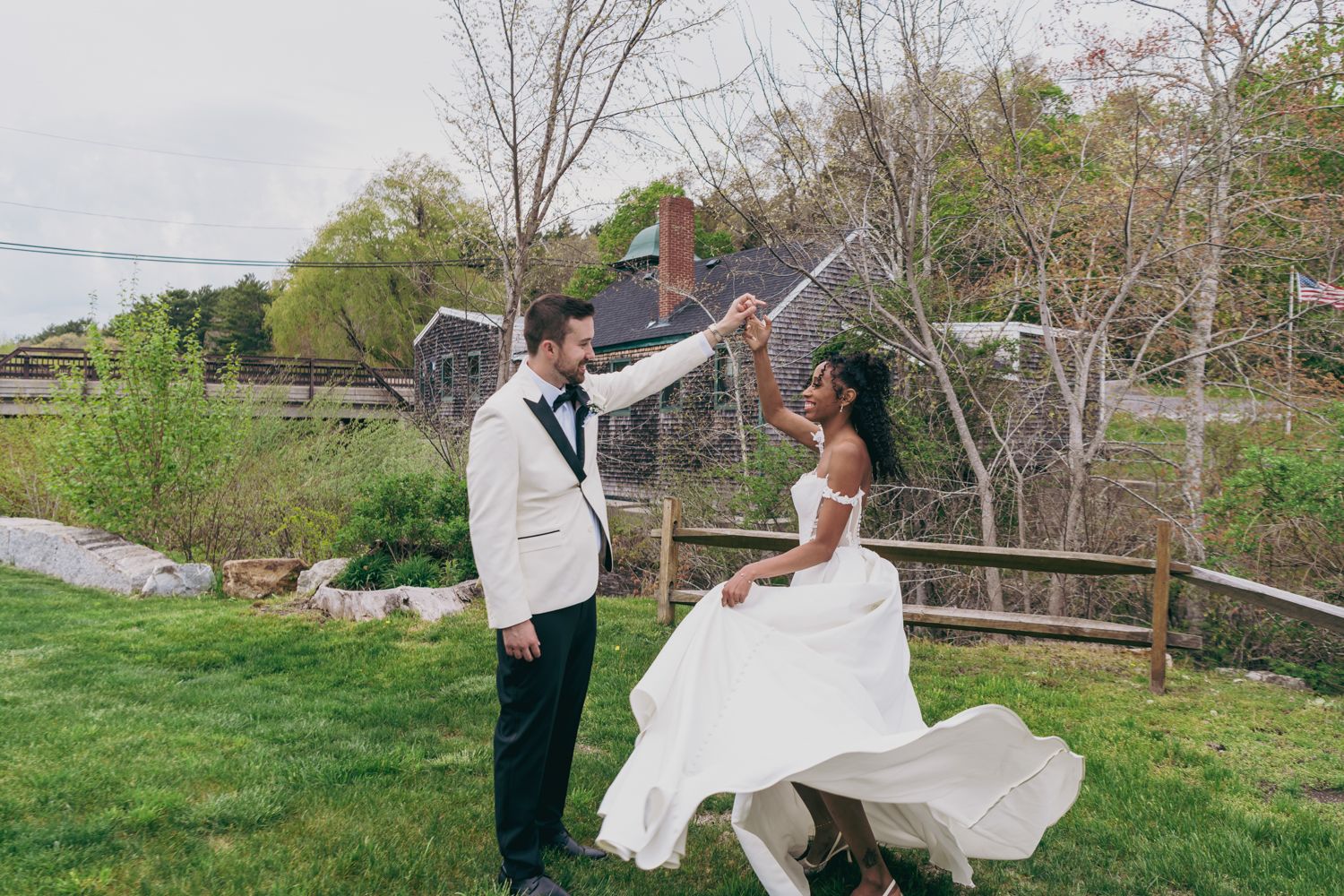 Groom twirling bride in flowing white wedding dress on green lawn outdoors.