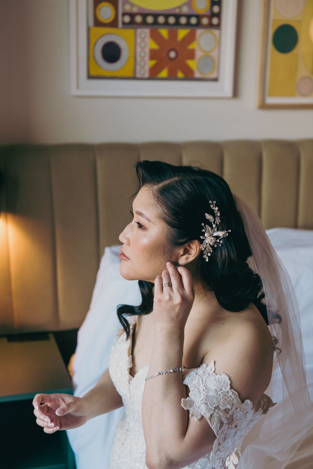 Bride in lace off-shoulder gown adjusting pearl earring, wearing crystal hair comb and veil, seated in elegant room.