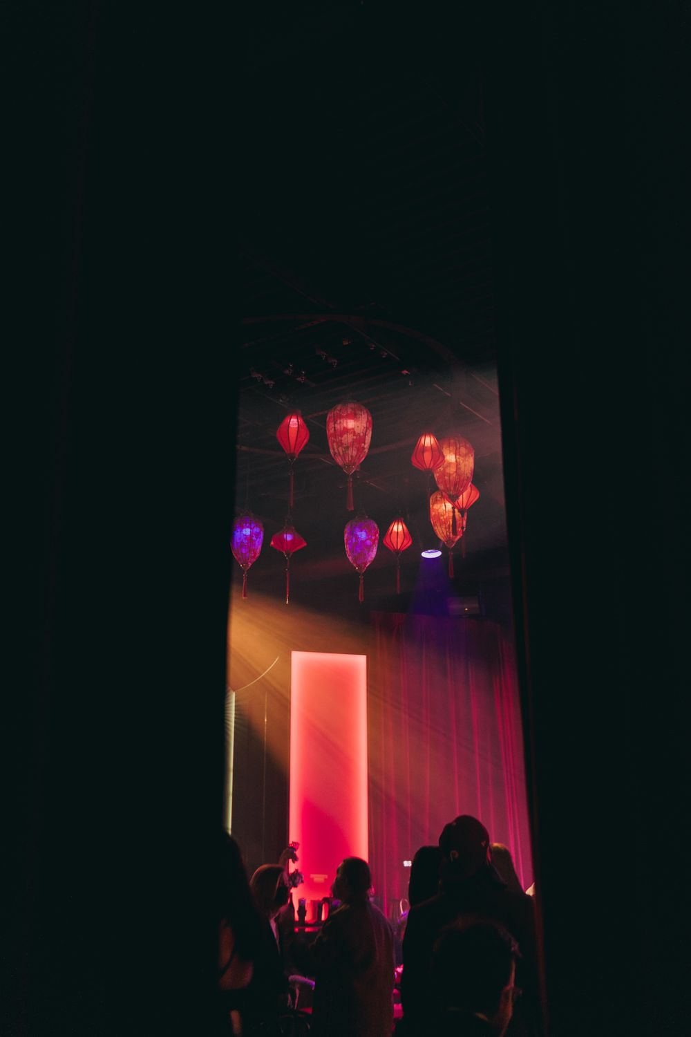 Dark wedding venue with glowing red and pink Chinese lanterns hanging from the ceiling above silhouetted guests.