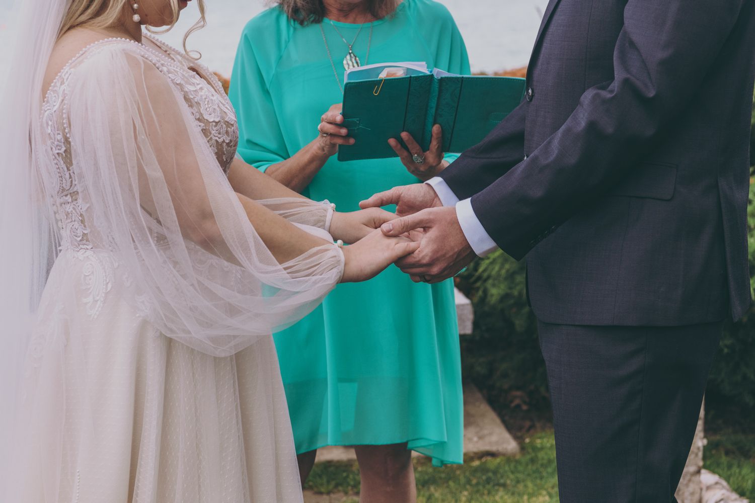 An officiant in teal reads vows as a bride and groom exchange rings during an outdoor ceremony.