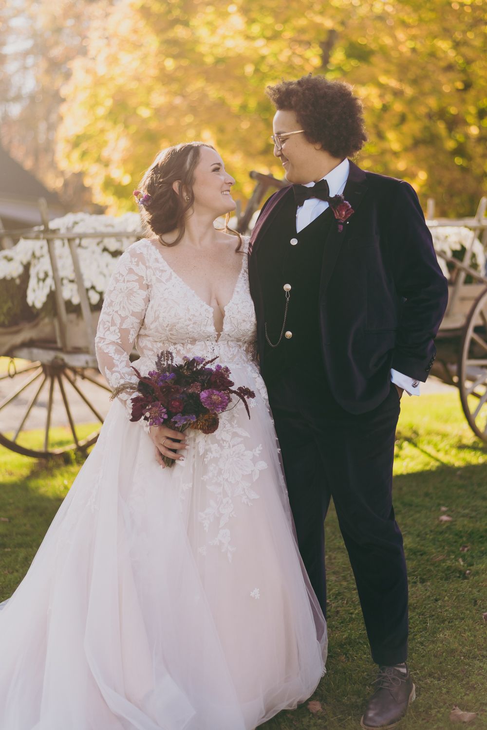 Bride and groom sharing a joyful moment in golden autumn sunlight beside a vintage wooden carriage.