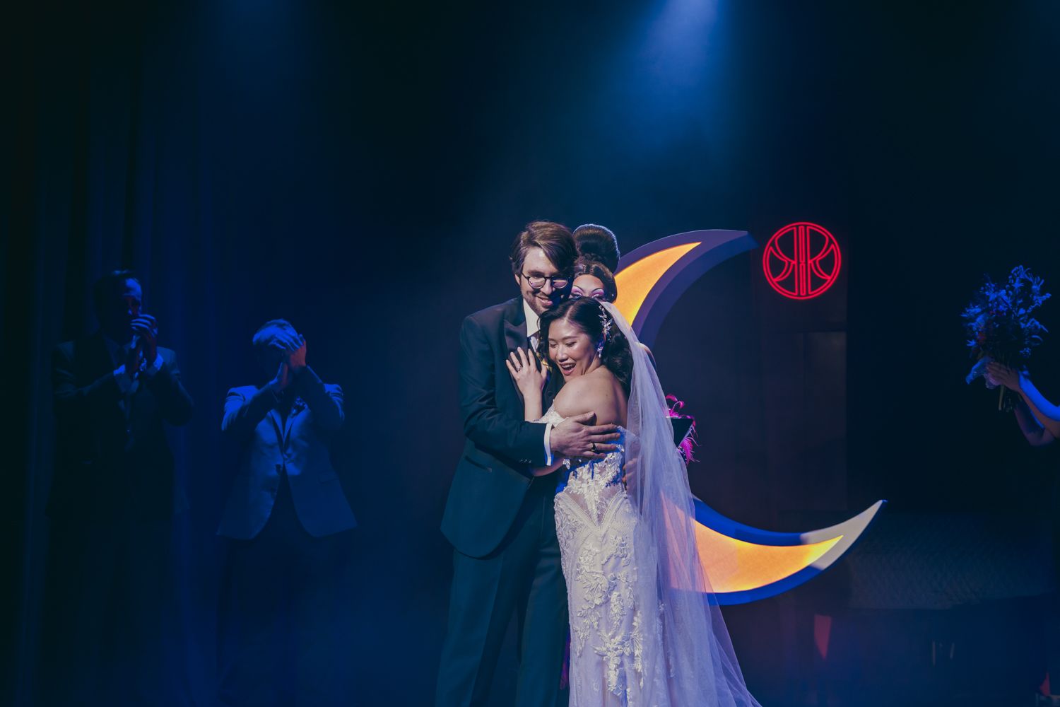 Bride and groom share romantic first dance on stage with crescent moon backdrop in dramatic blue lighting.