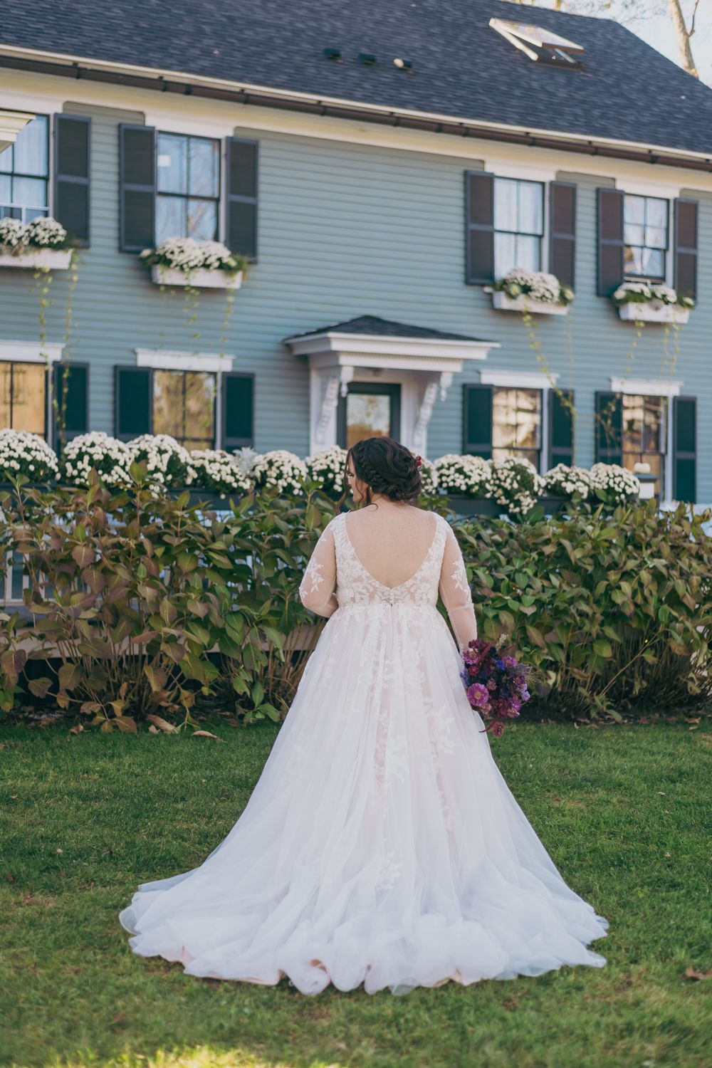 Bride in elegant lace wedding gown with low back holding purple bouquet standing before a classic New England inn.