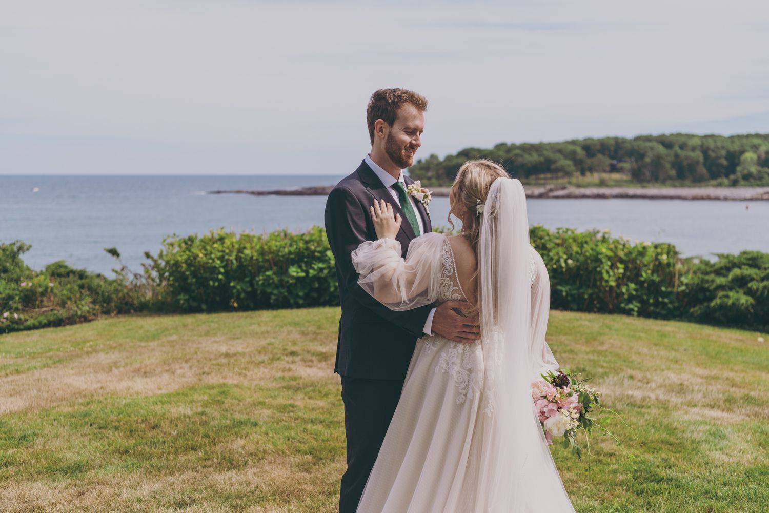 Bride and groom share a romantic embrace on a green lawn overlooking a scenic coastal bay on their wedding day.