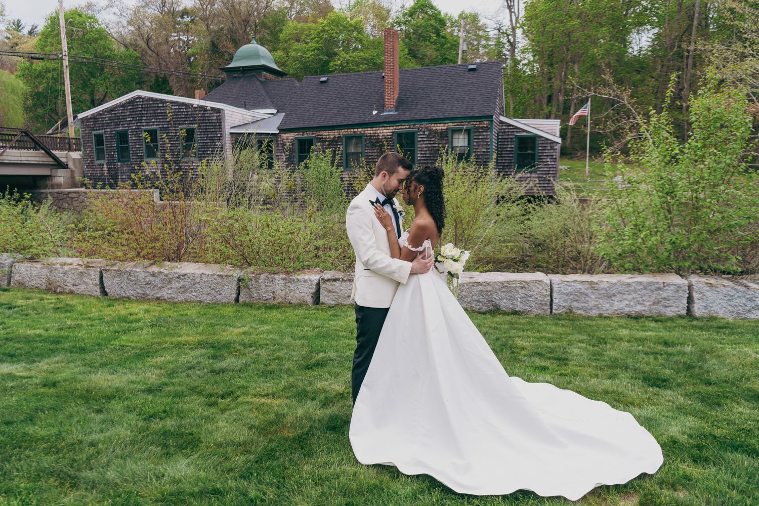 Interracial couple sharing a romantic kiss on green lawn with rustic building backdrop on their wedding day.