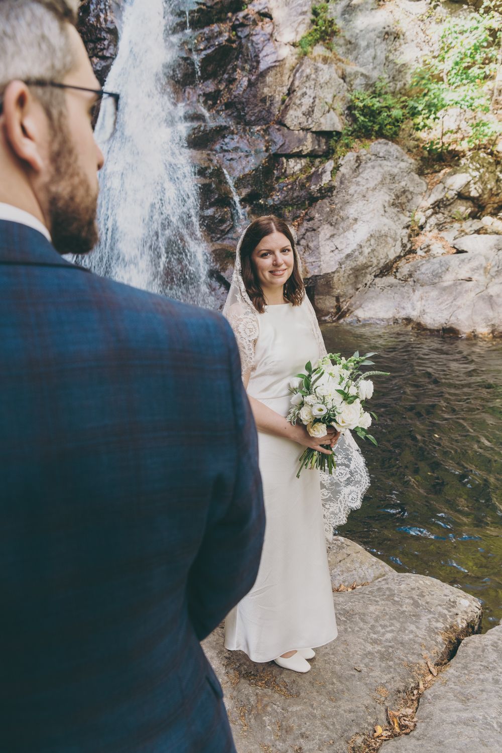 Smiling bride holds white bouquet while groom faces her before a stunning waterfall.