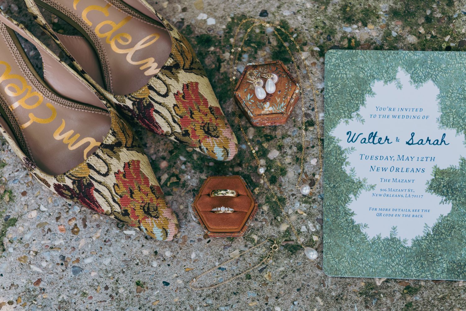 Wedding flat lay featuring floral heels, ring boxes, gold rings, and a green Walter and Sarah invitation.