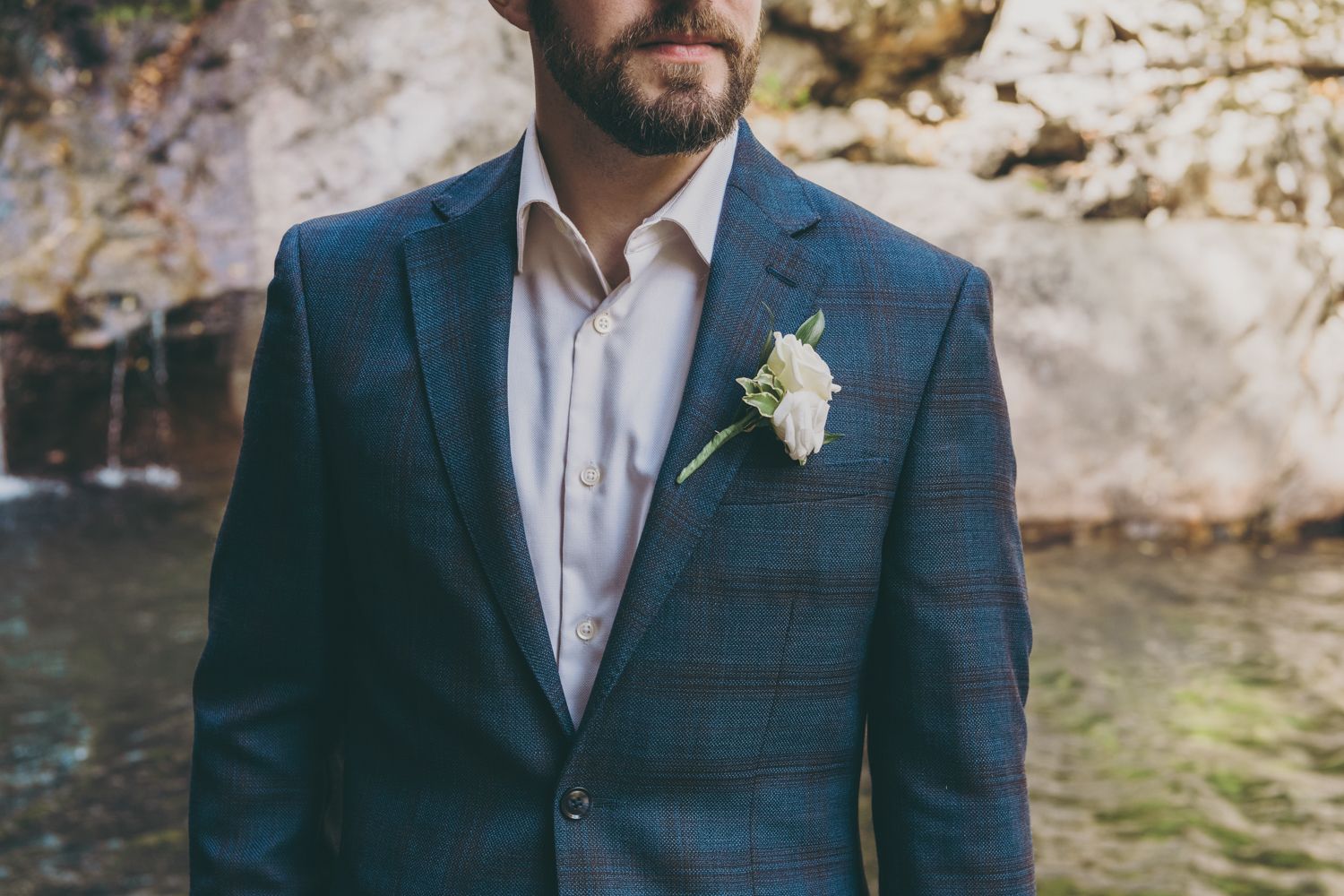 Dapper groom in plaid blue suit with white boutonniere poses near rocky waterfall.