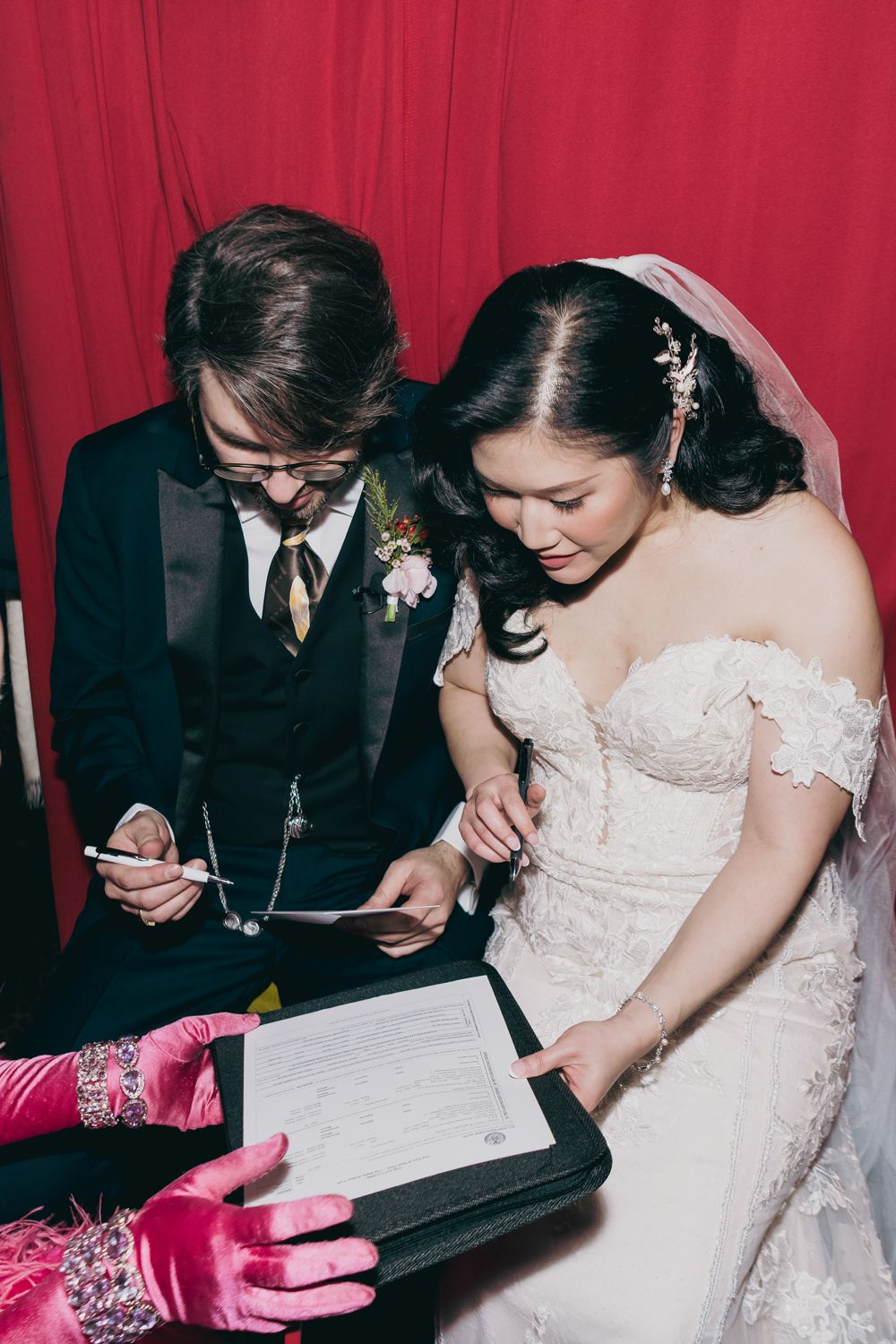 Bride and groom sign wedding certificate together, bride in lace off-shoulder gown with veil, against red curtain backdrop.