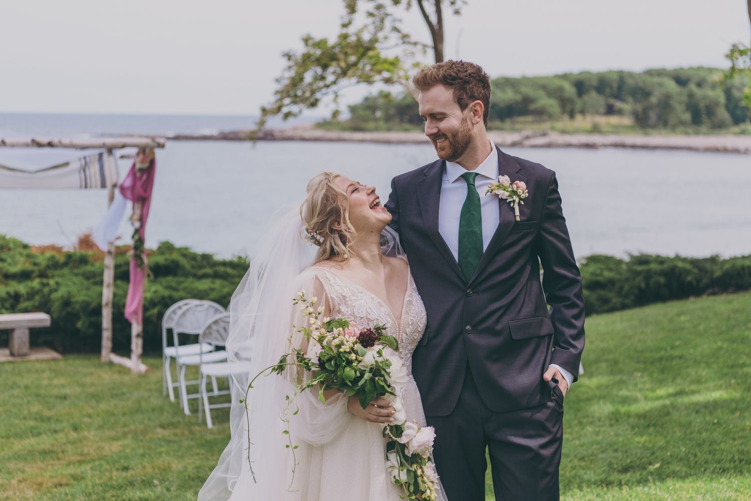 A smiling bride and groom walk together outdoors near a waterfront wedding ceremony backdrop.