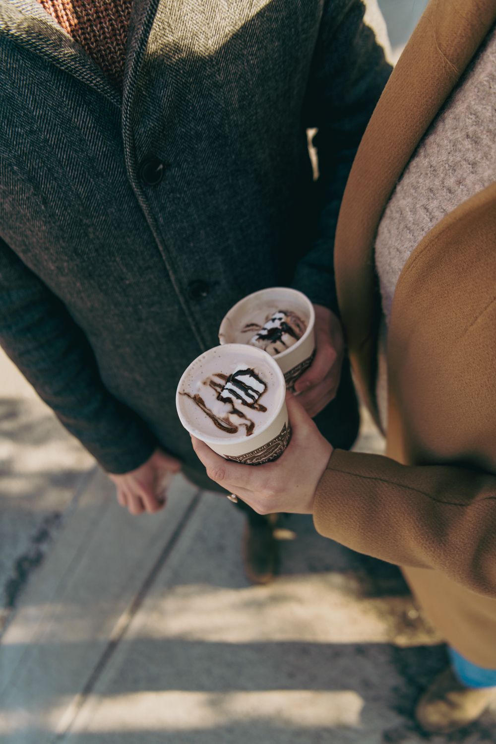 Couple holding two takeaway coffee cups with chocolate drizzle on top while standing outdoors in autumn coats.