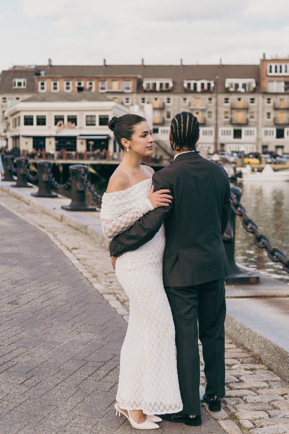 Interracial couple embracing on a waterfront promenade, bride in white off-shoulder gown and groom in dark suit.