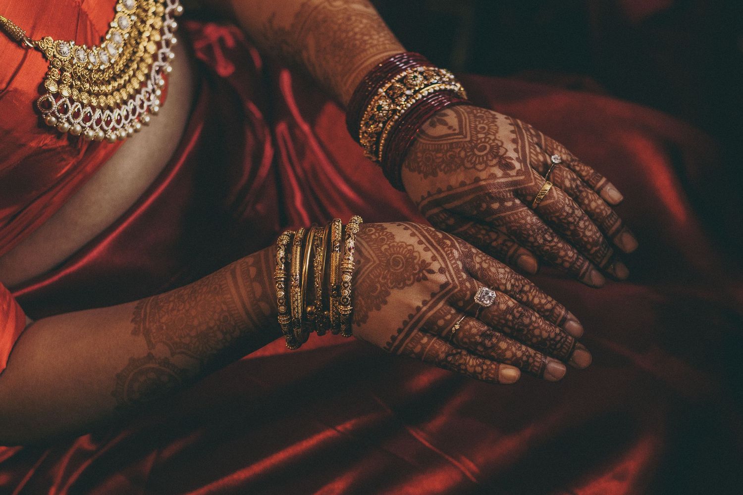 Close-up of Indian bride's hands adorned with intricate henna mehndi patterns and gold bangles on red silk sari.
