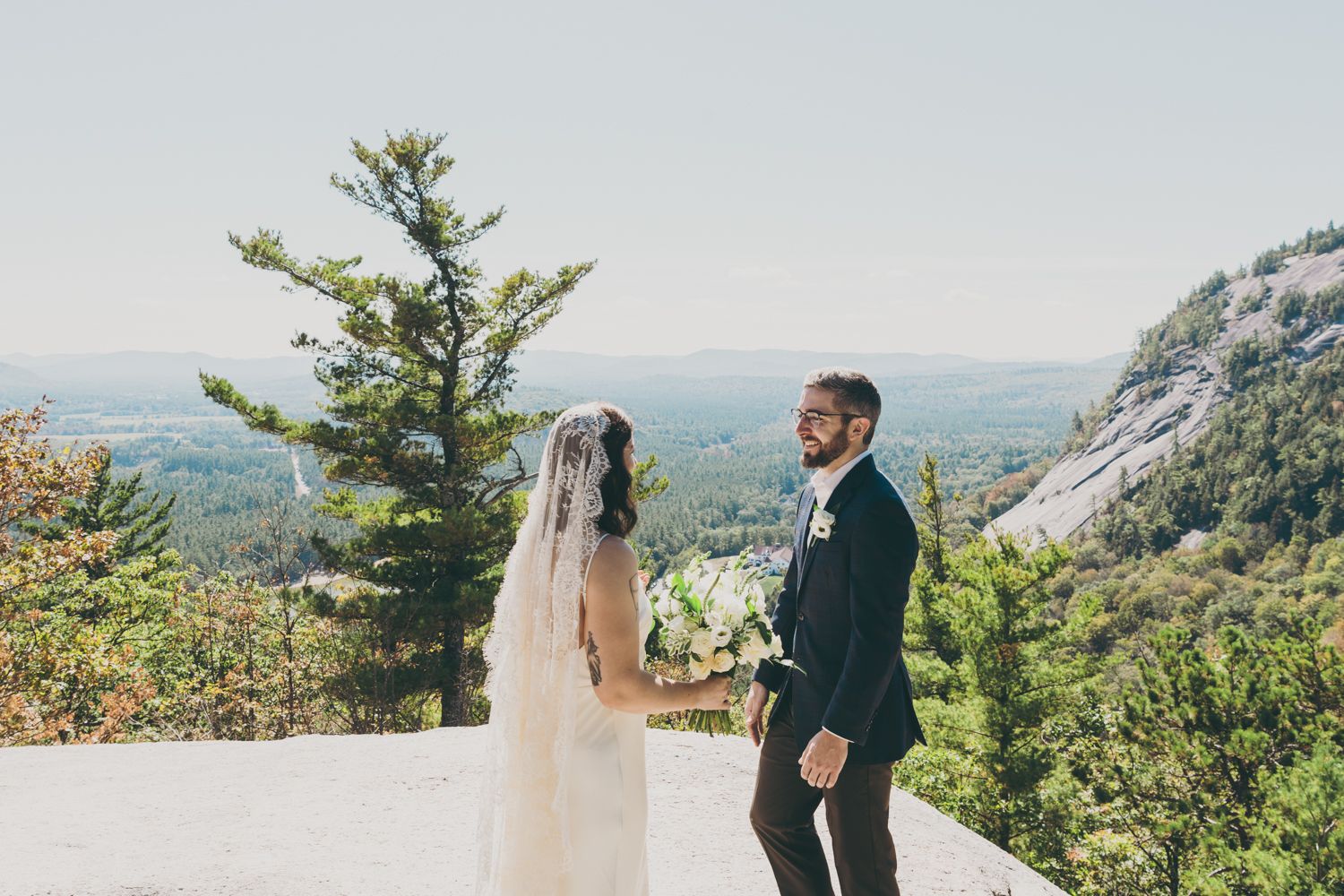 Groom smiling joyfully as bride in white veil turns around during outdoor mountain first look.
